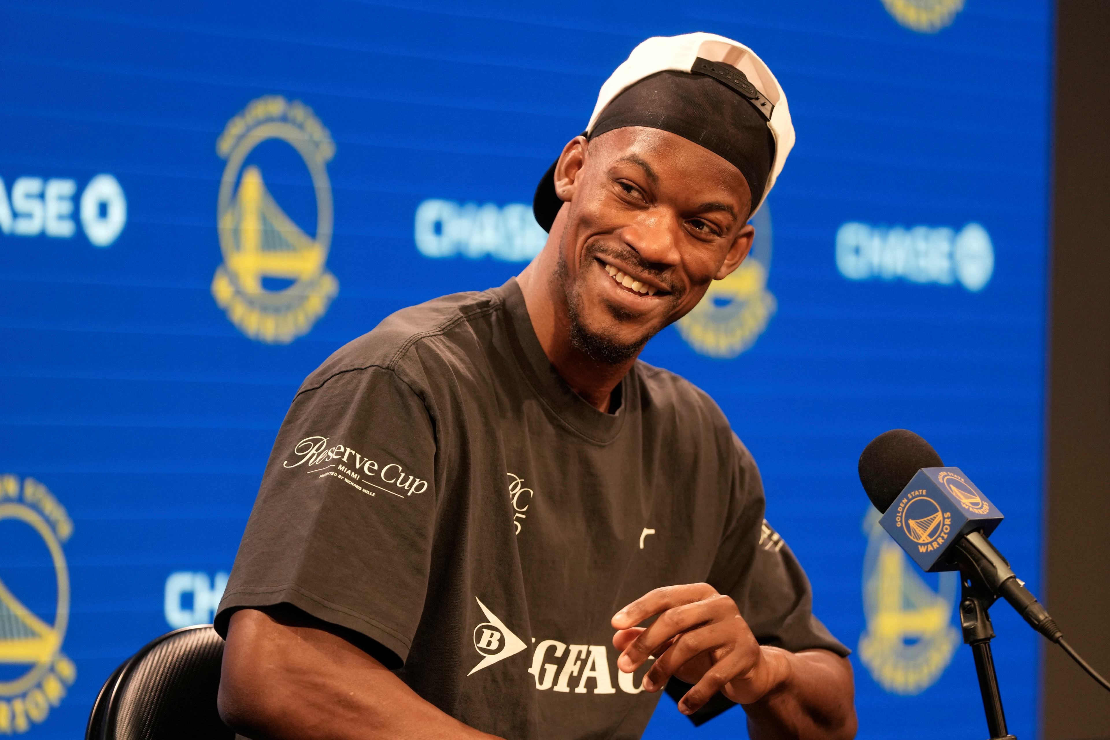 A man in a black shirt and backwards cap smiles at a press event with a Golden State Warriors backdrop. A microphone is in front of him.