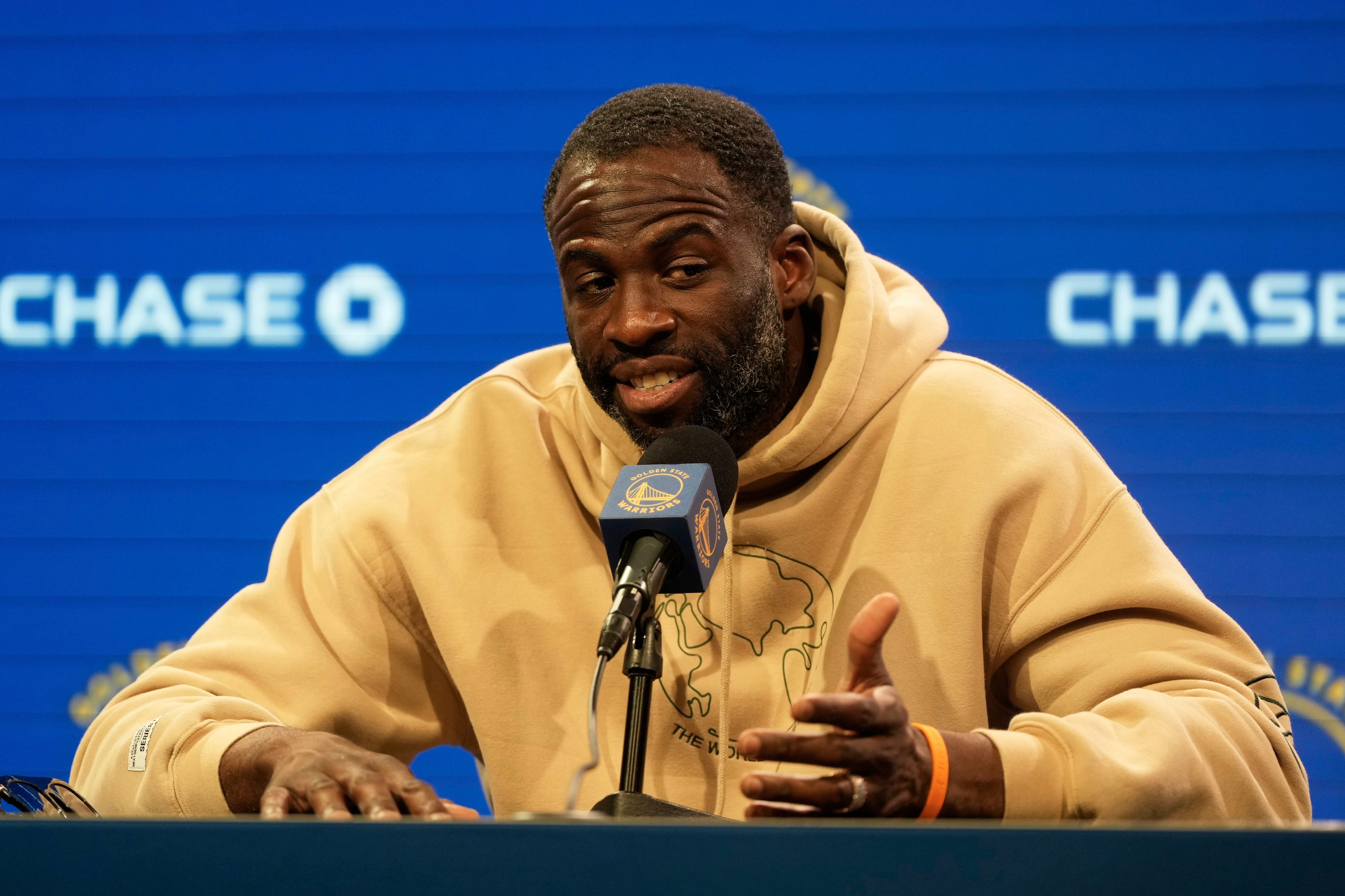 A man in a beige hoodie speaks into a microphone at a press conference. The backdrop is blue with &quot;CHASE&quot; logos.