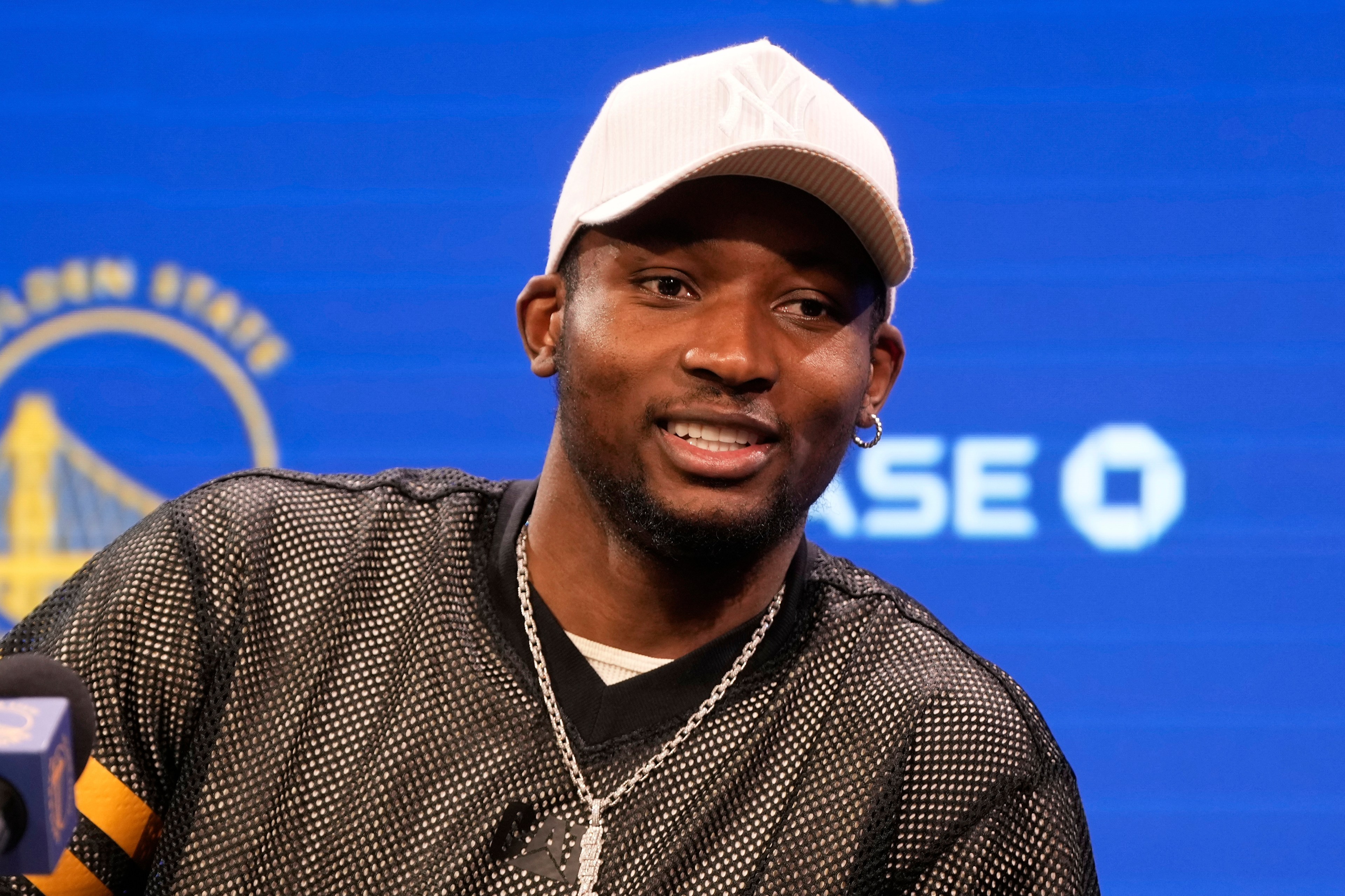A person is smiling, wearing a white cap and a mesh shirt with a necklace. The background is blue with a Golden State Warriors logo.