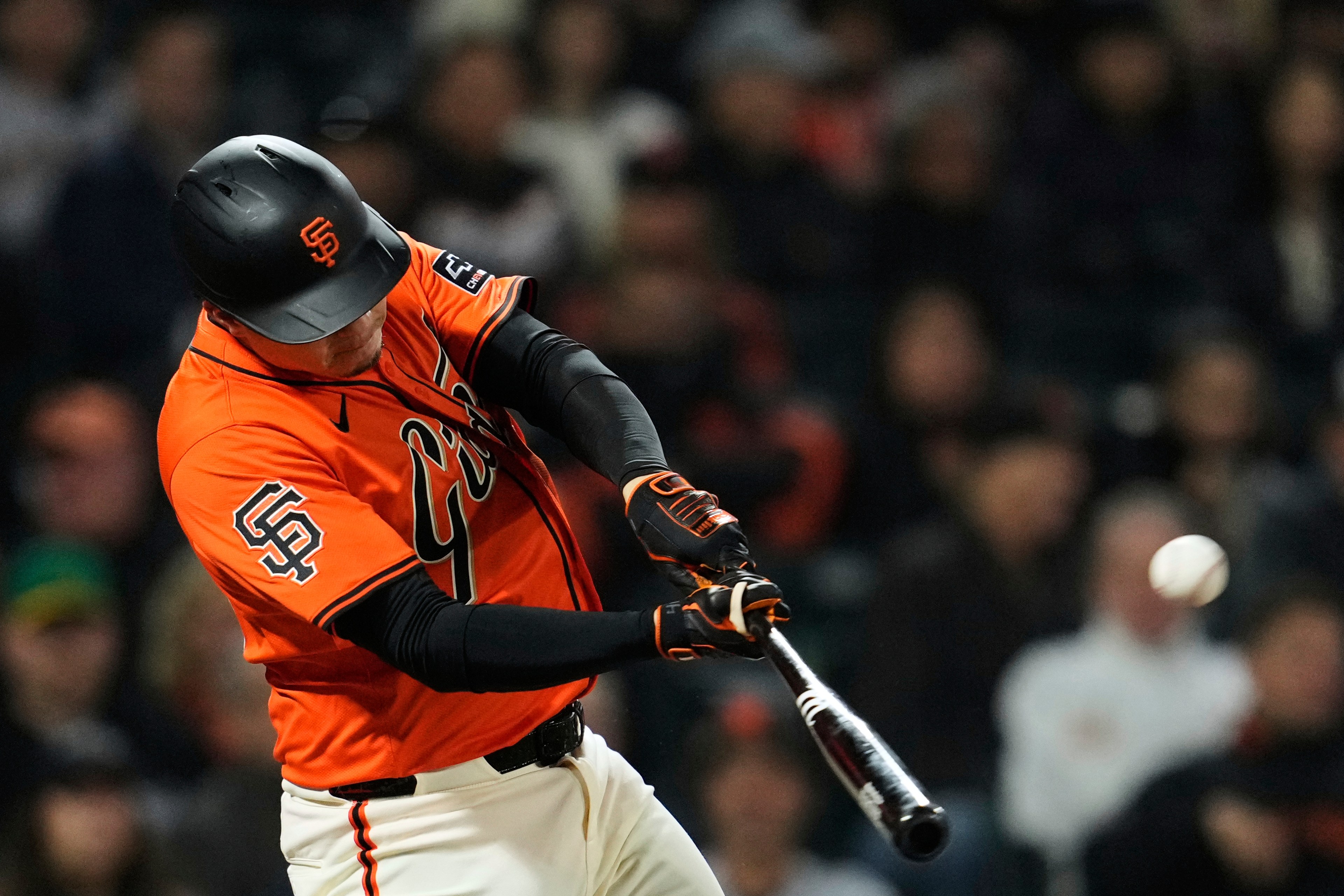 A baseball player in an orange jersey and helmet swings a bat, just making contact with the ball. The background is blurred with spectators.