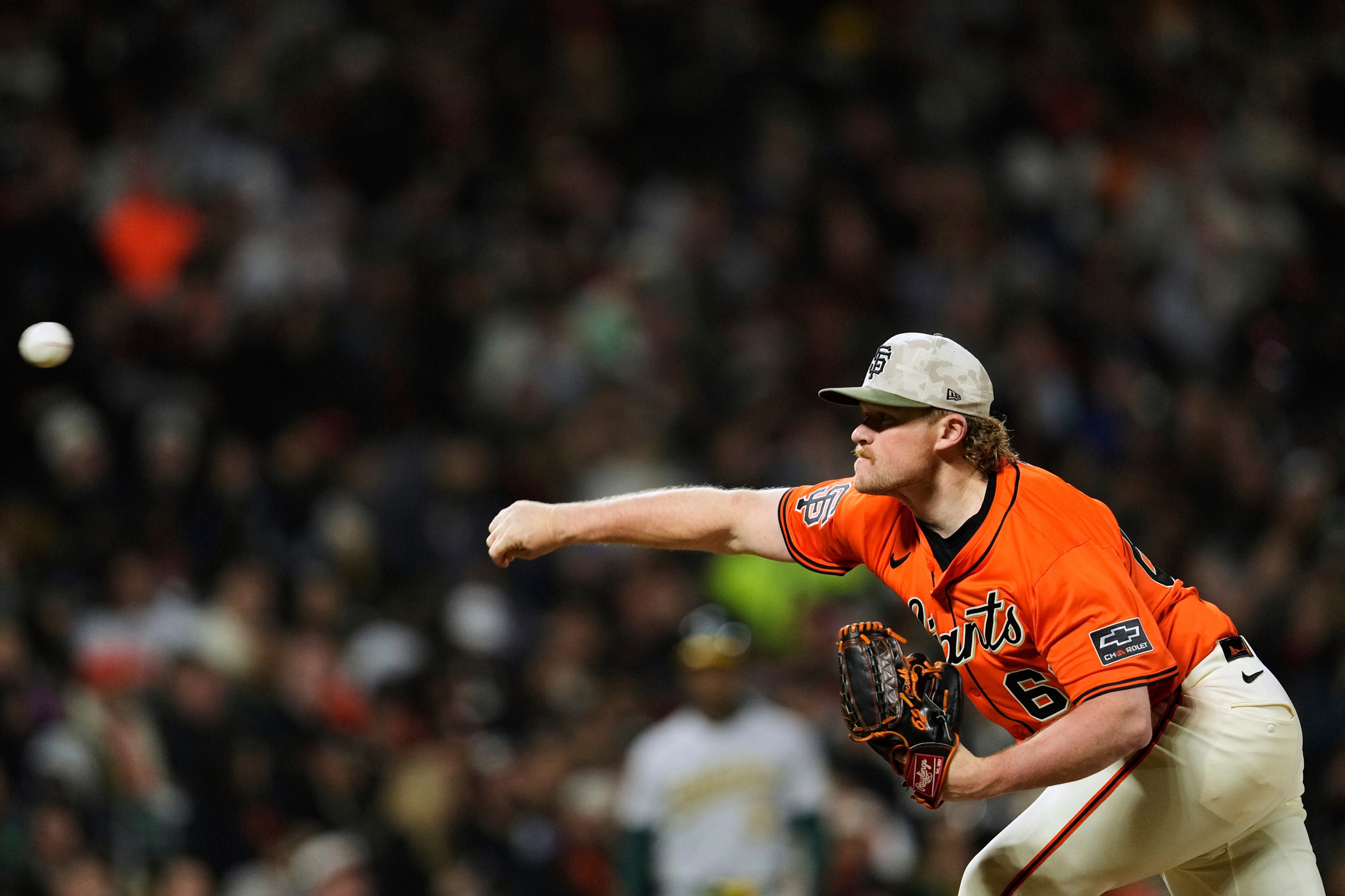 A baseball player in an orange jersey and cap is pitching the ball. The crowd in the background is blurred, and a batsman stands in focus.