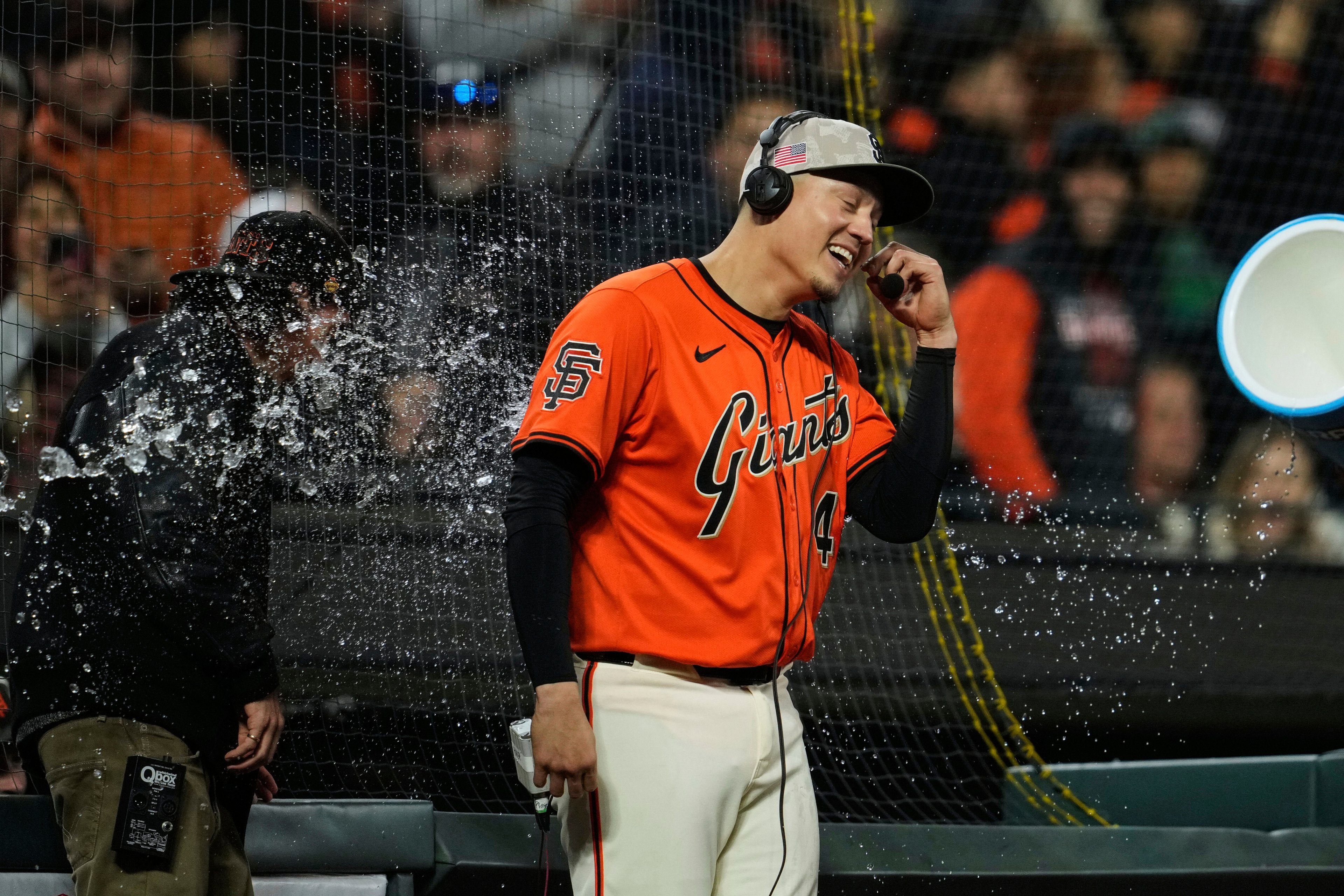 A baseball player in a Giants uniform cheerfully wears a headset while water is splashed over another person beside him, with a large bucket visible in the air.