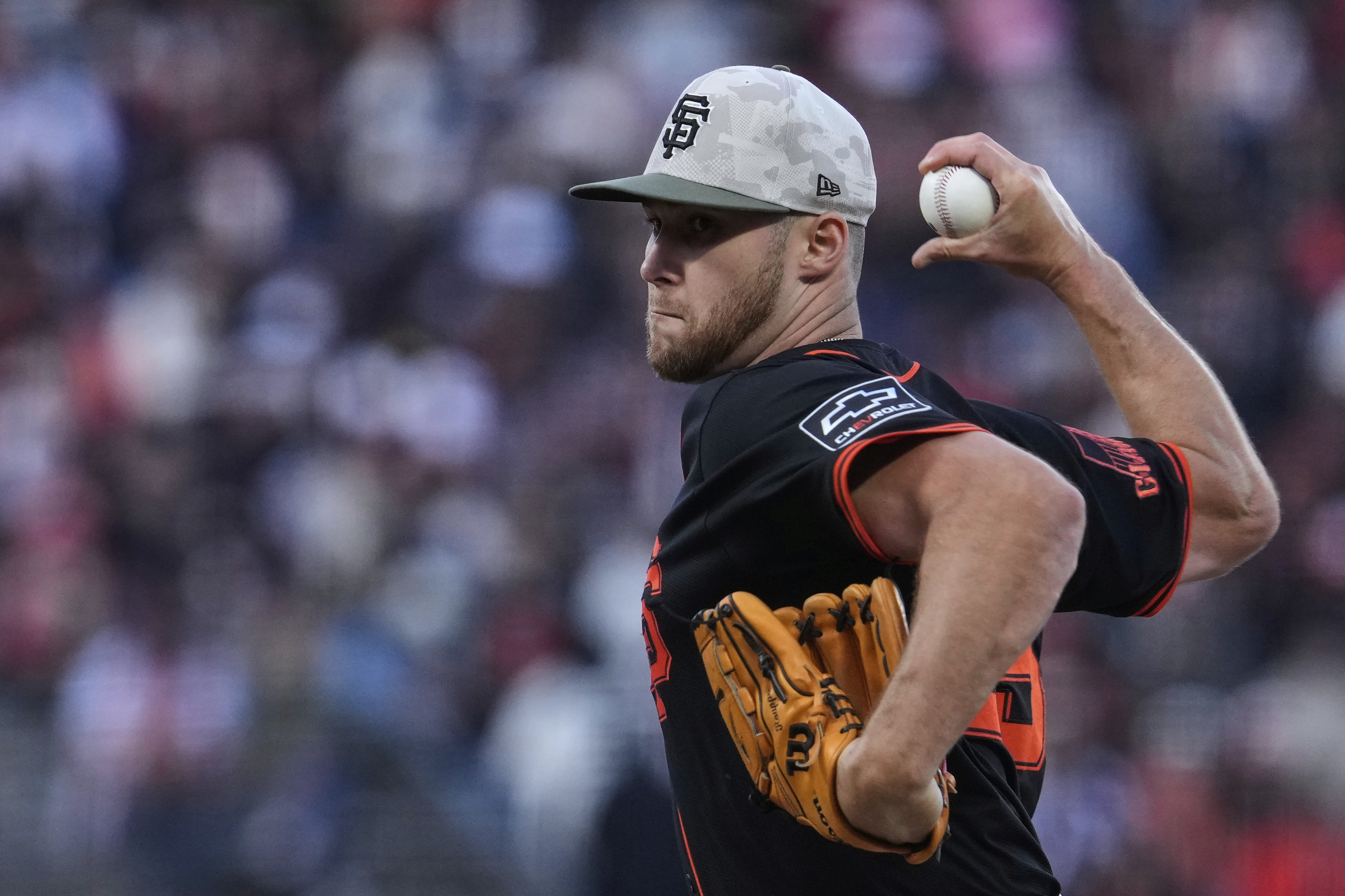 A baseball player in a black uniform and camo cap is pitching, gripping a baseball. He wears an orange glove and has a focused expression.