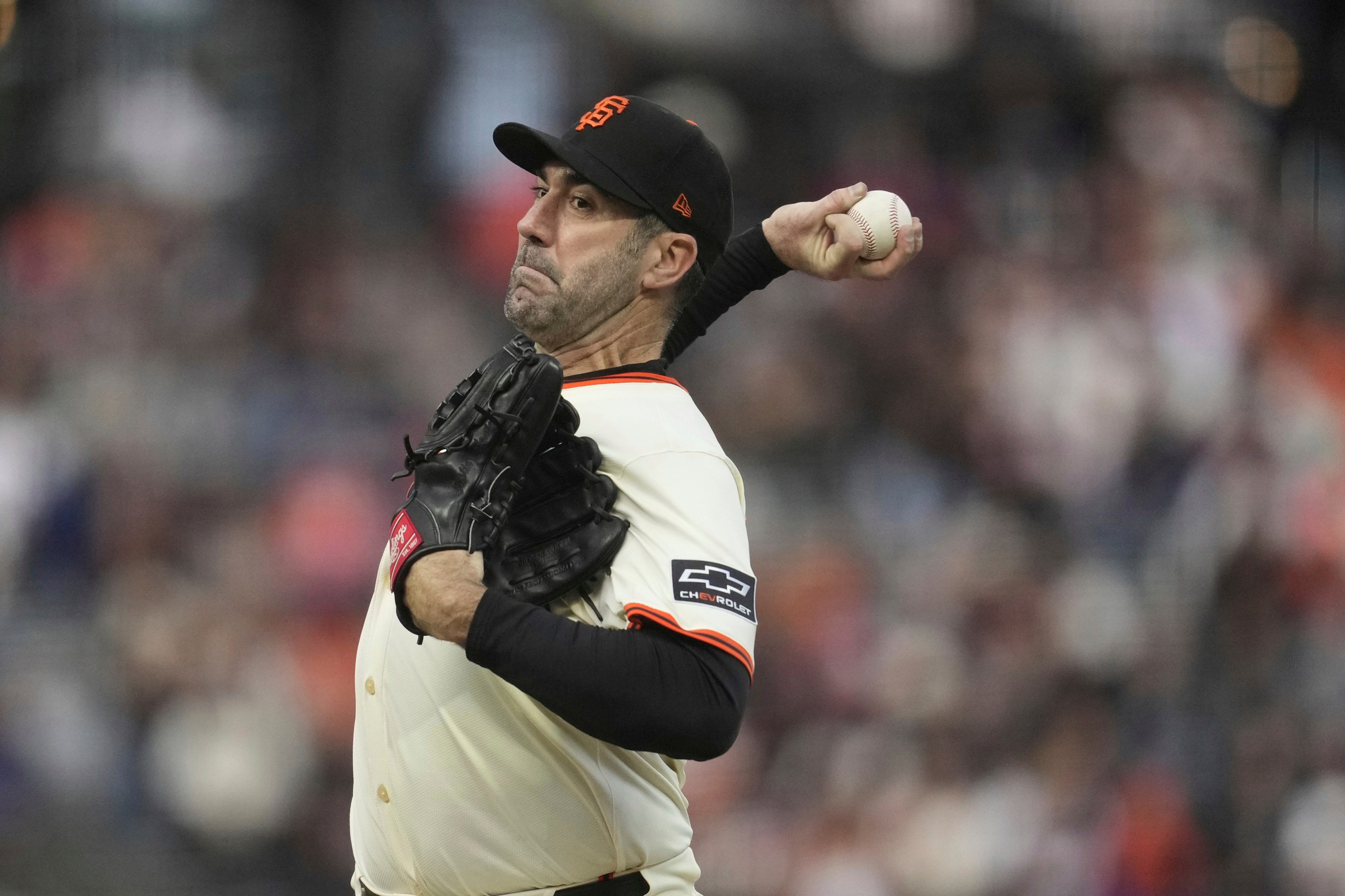 A baseball player in a white uniform with black trim and an orange "SF" cap is mid-pitch, holding a baseball in one hand and wearing a black glove on the other.