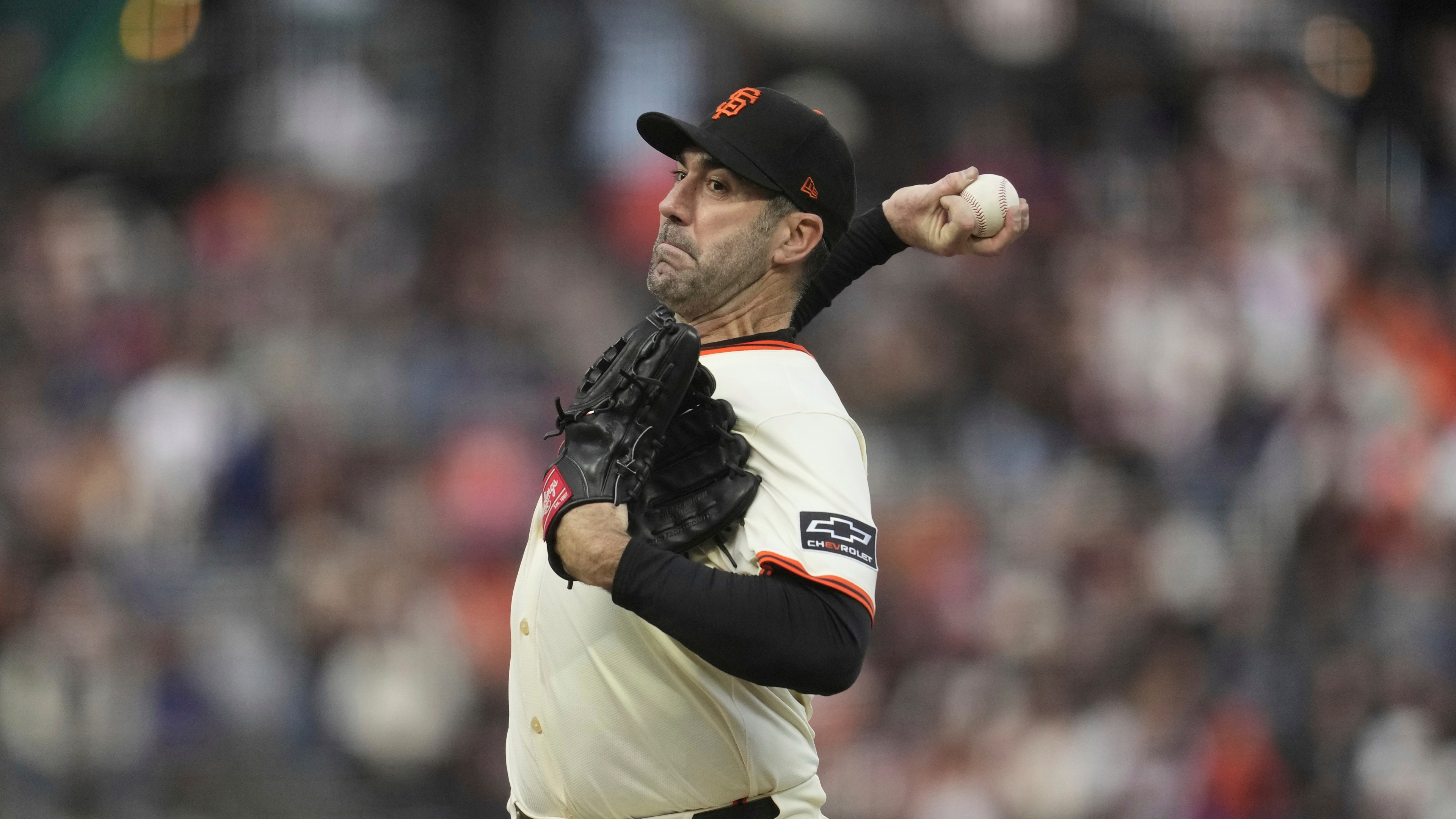 A baseball player in a white uniform with black trim and an orange "SF" cap is mid-pitch, holding a baseball in one hand and wearing a black glove on the other.
