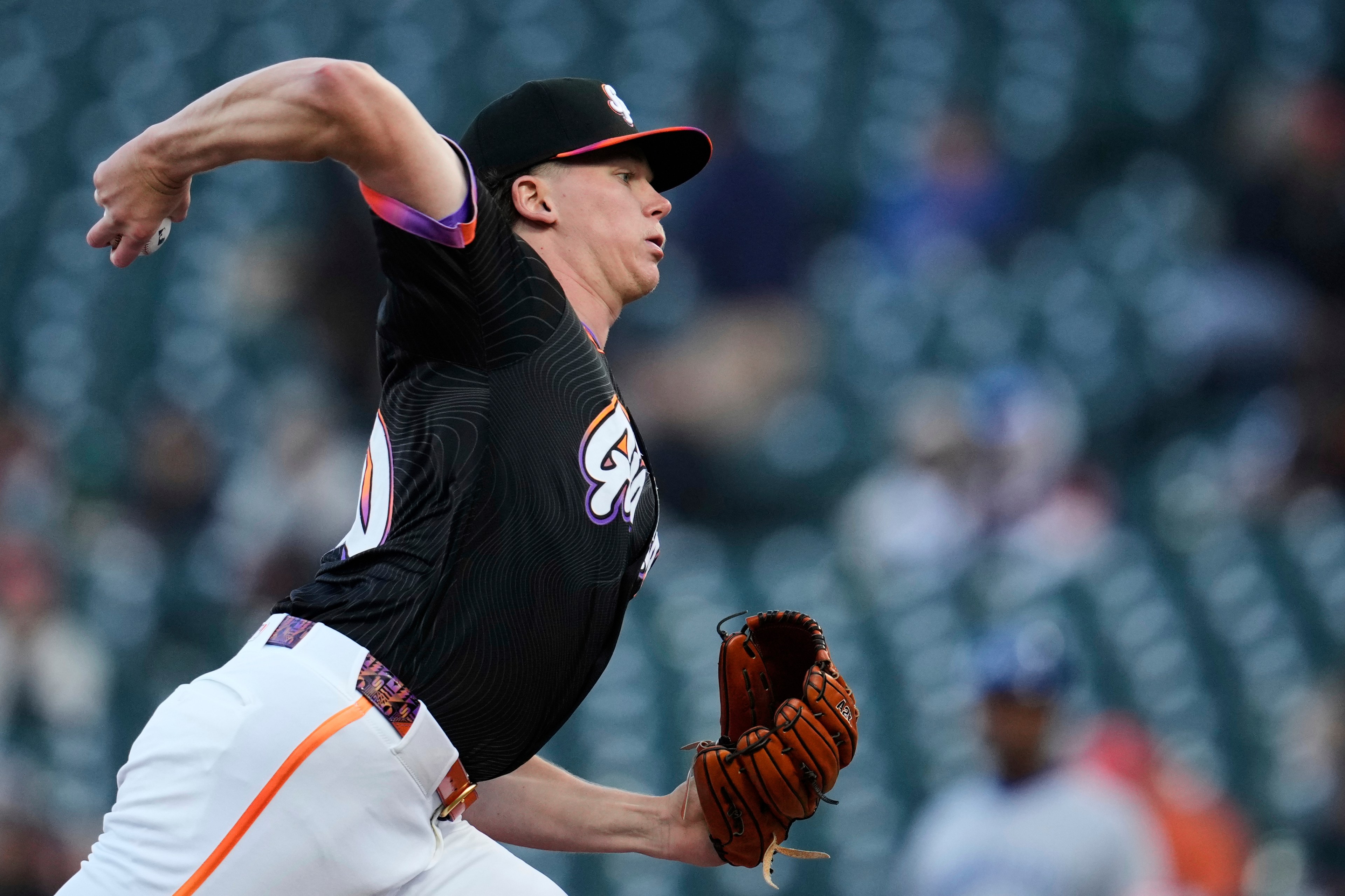 A baseball player in a black jersey is pitching, with his right arm extended and a glove in his left hand. He is in a stadium with empty green seats behind him.
