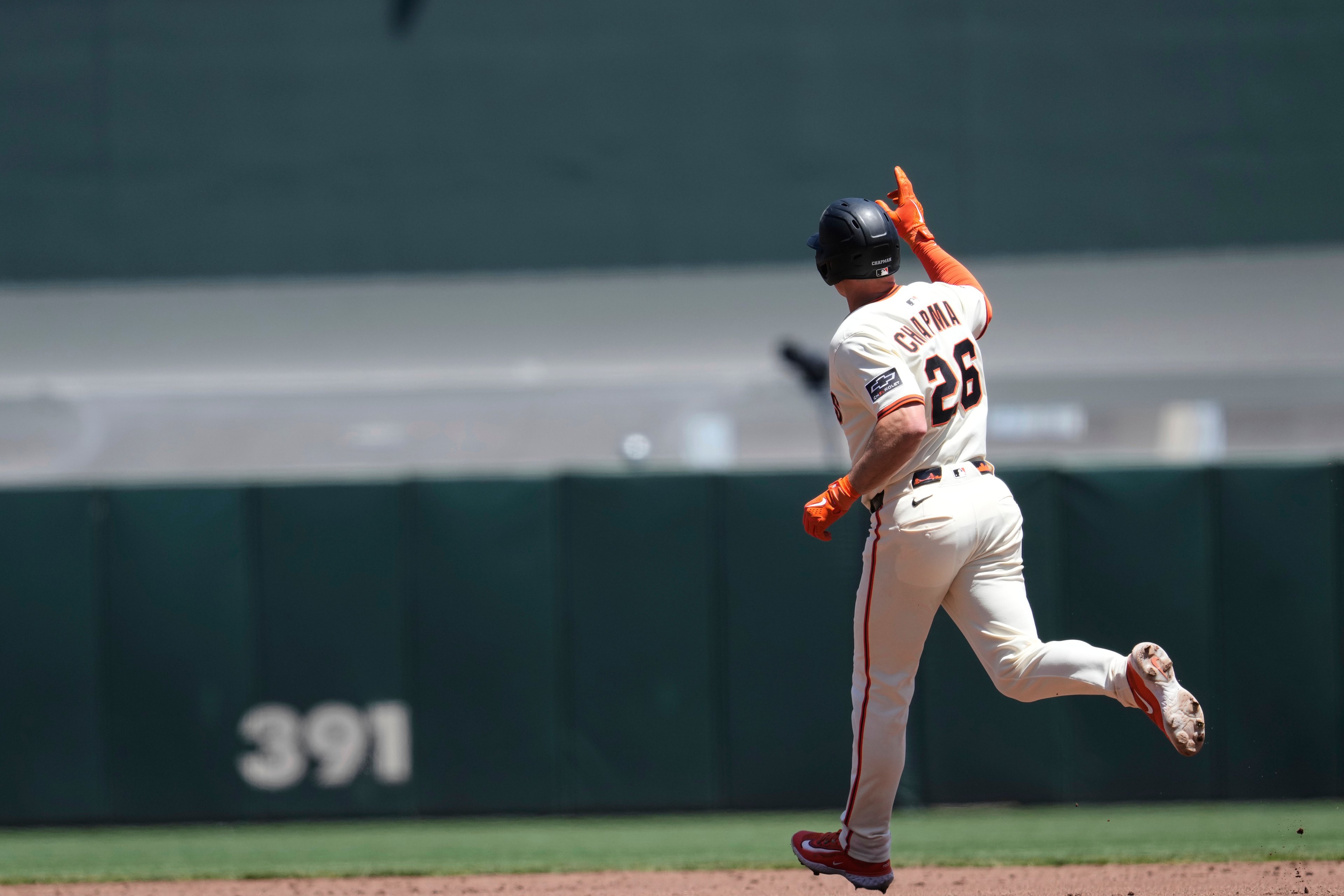 A baseball player in a white uniform runs on the field, pointing upwards with one hand. His jersey has the number 26, and the scoreboard reads &quot;391&quot; in the background.