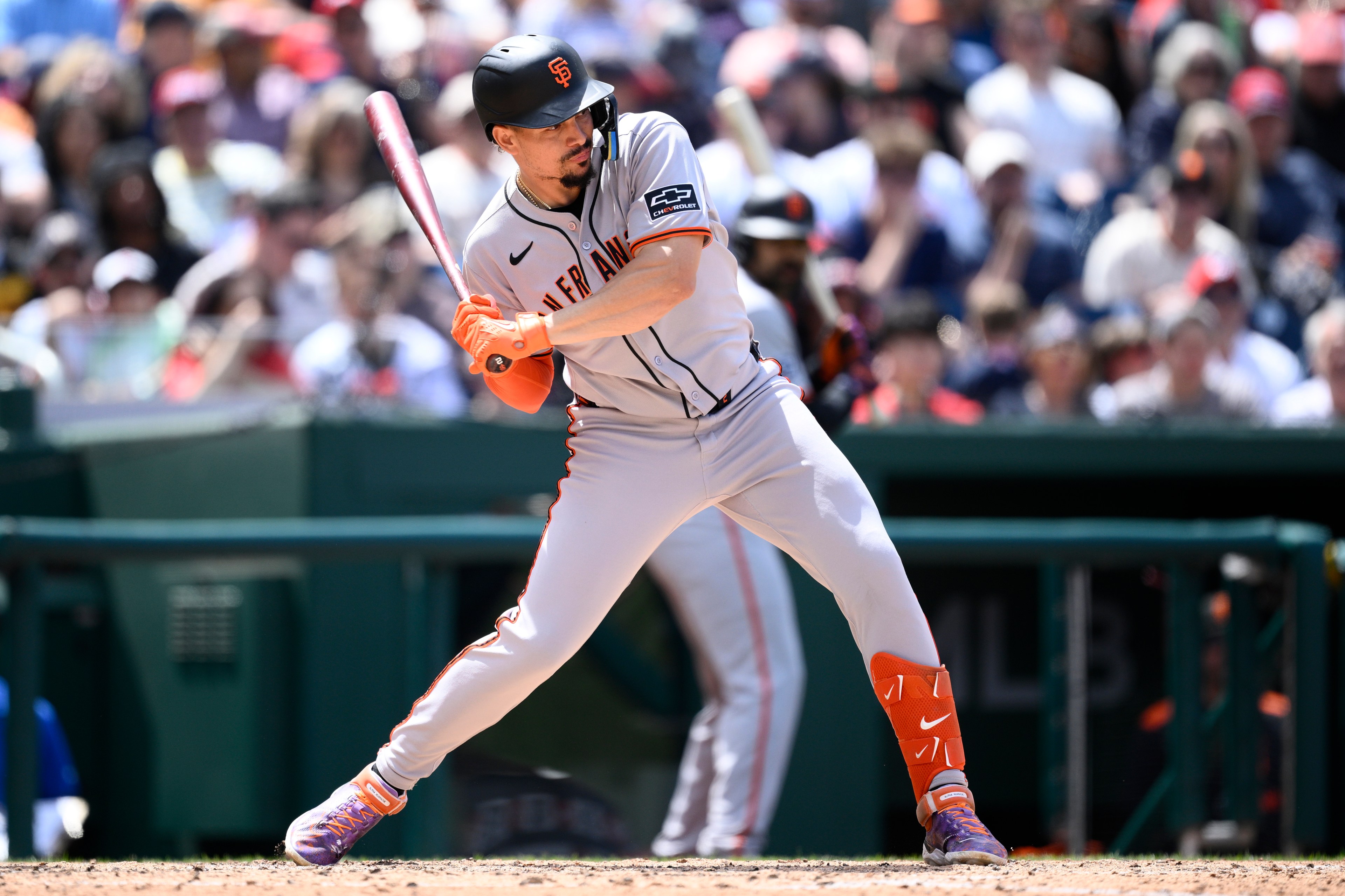 A baseball player in a gray and orange uniform with a black helmet is swinging a bat. The background shows a crowd watching attentively.