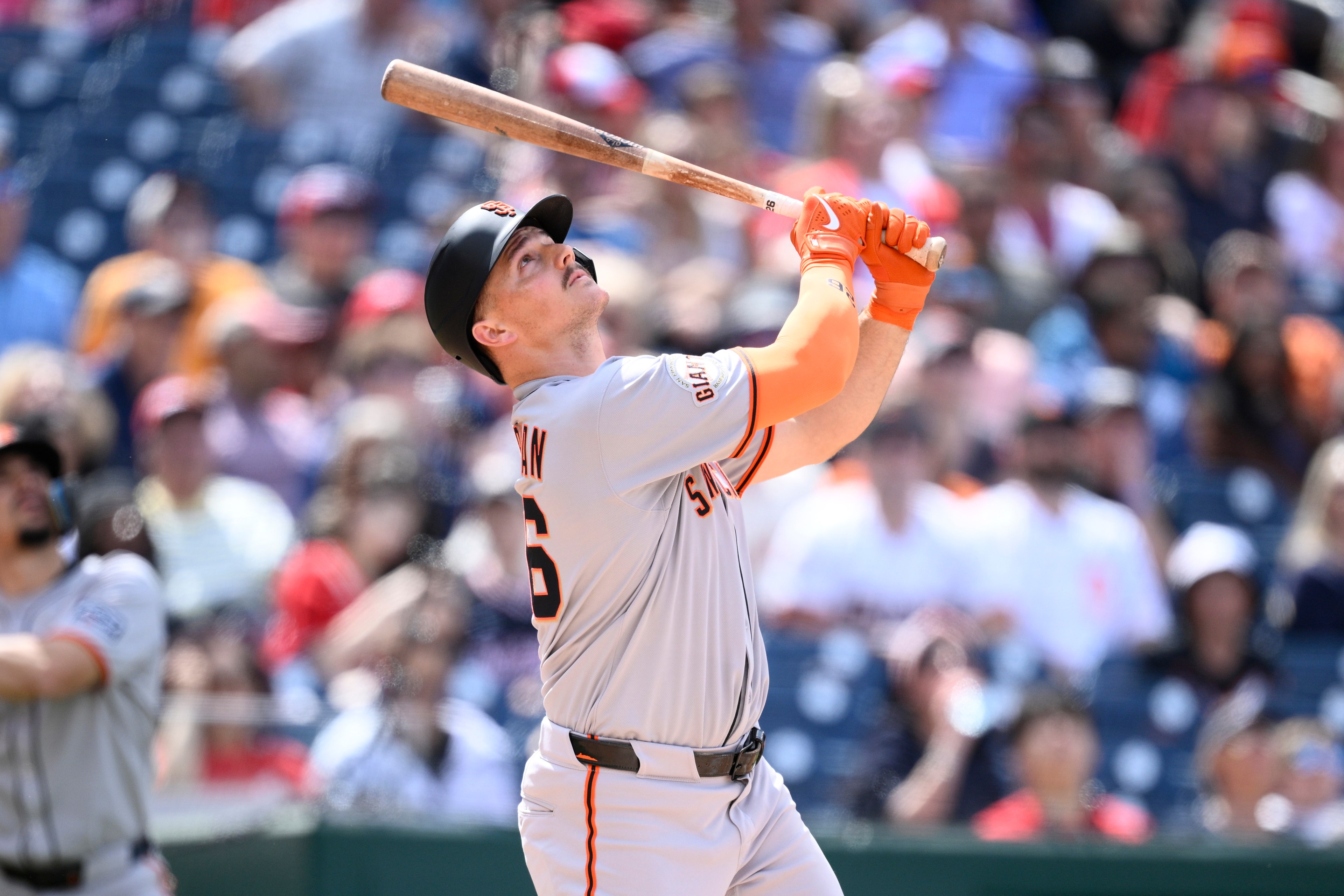A baseball player in a gray uniform and black helmet watches his hit while holding a bat. The crowd is blurred in the background, suggesting a sunny day at a game.