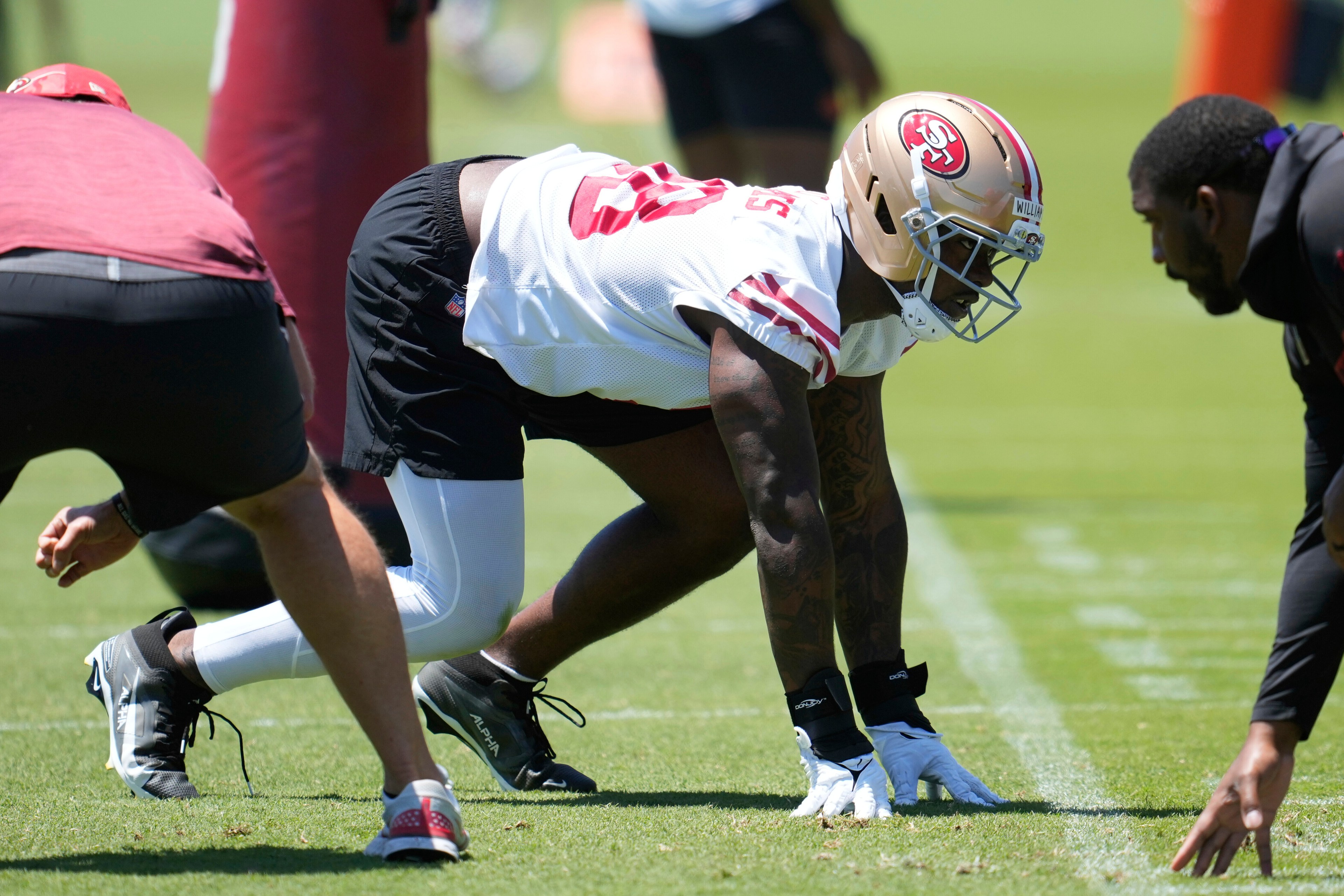 Two football players face each other crouched in a stance on a practice field, wearing helmets and team gear under a sunny sky.