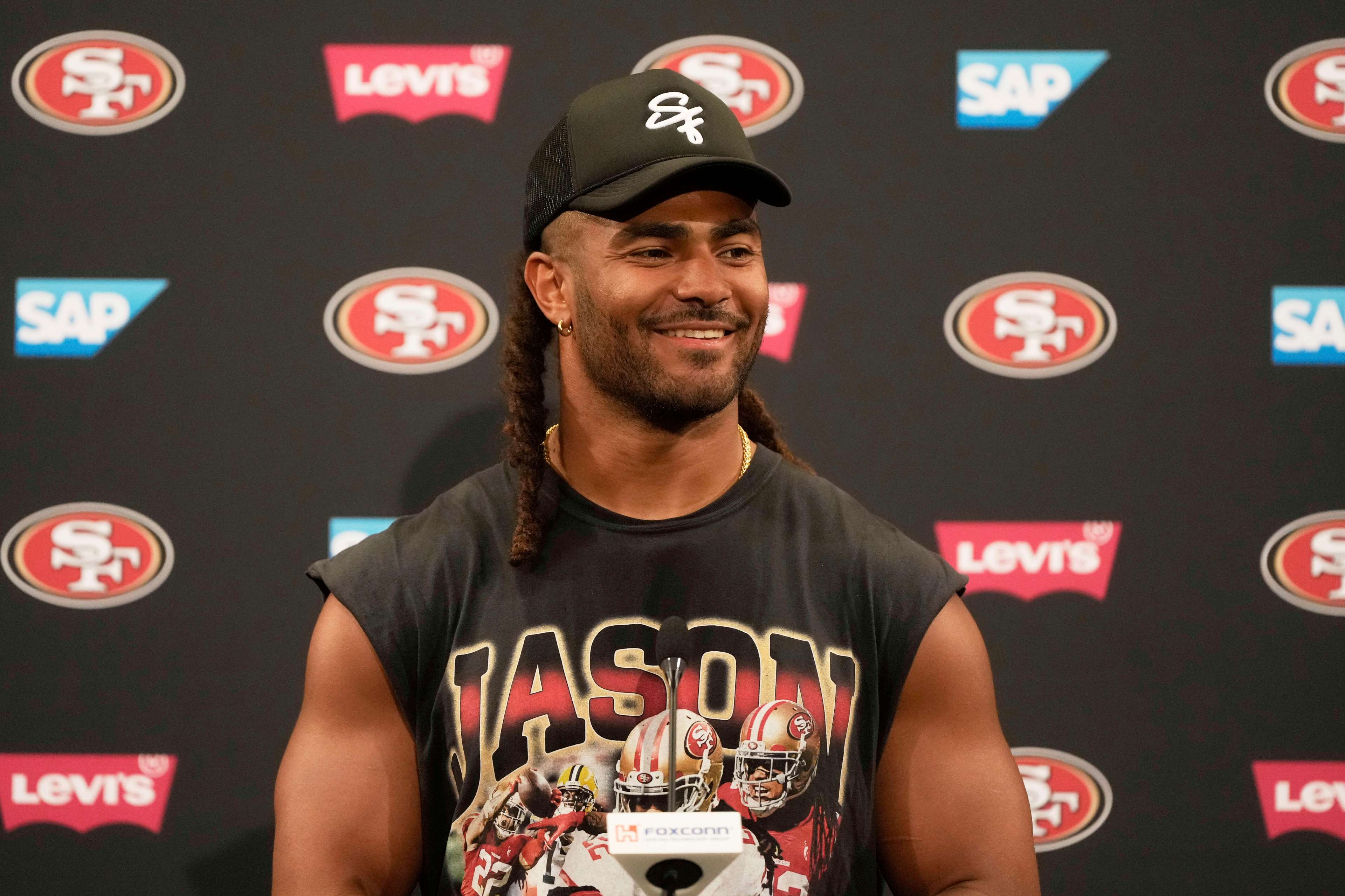 A person with long hair smiles at a press conference, wearing a black cap and a sleeveless shirt featuring football players. The backdrop has SAP and Levi’s logos.
