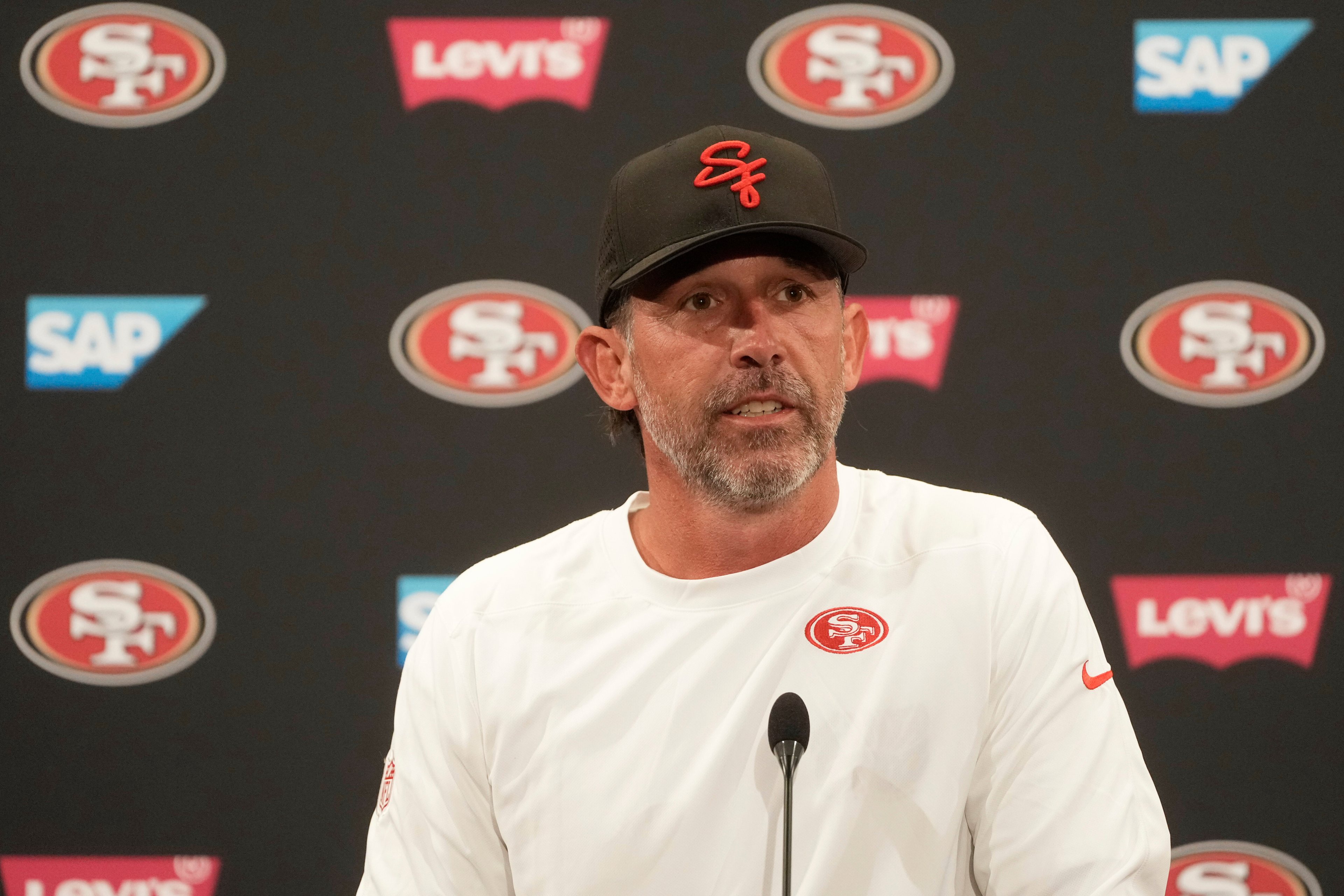 A man in a white shirt and black cap with red letters stands in front of a backdrop with logos, including &quot;49ers,&quot; SAP, and Levi's, speaking into a microphone.