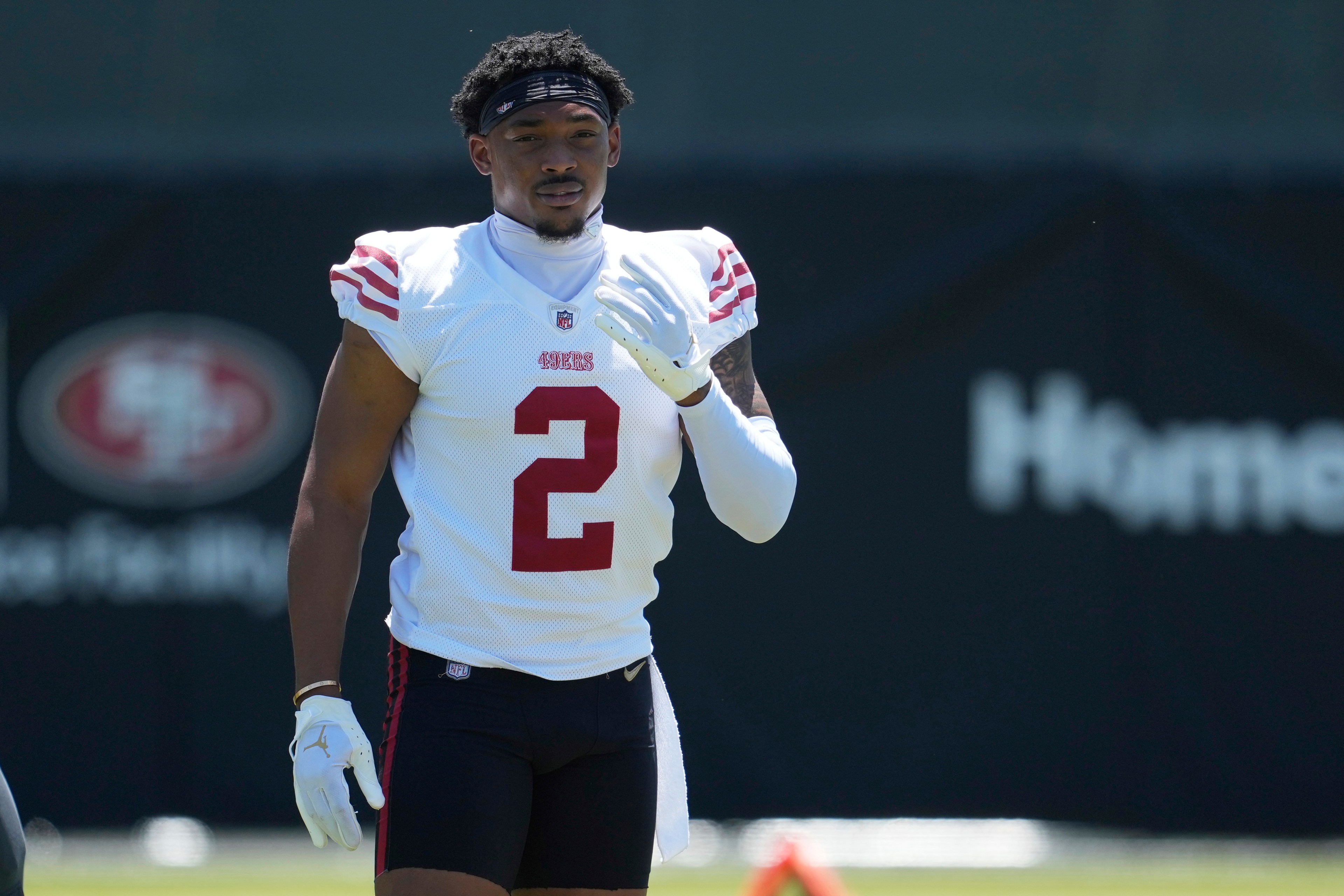 A football player in a white jersey with red stripes and the number 2 is on a field. He wears white gloves and a dark headband, walking towards the camera.