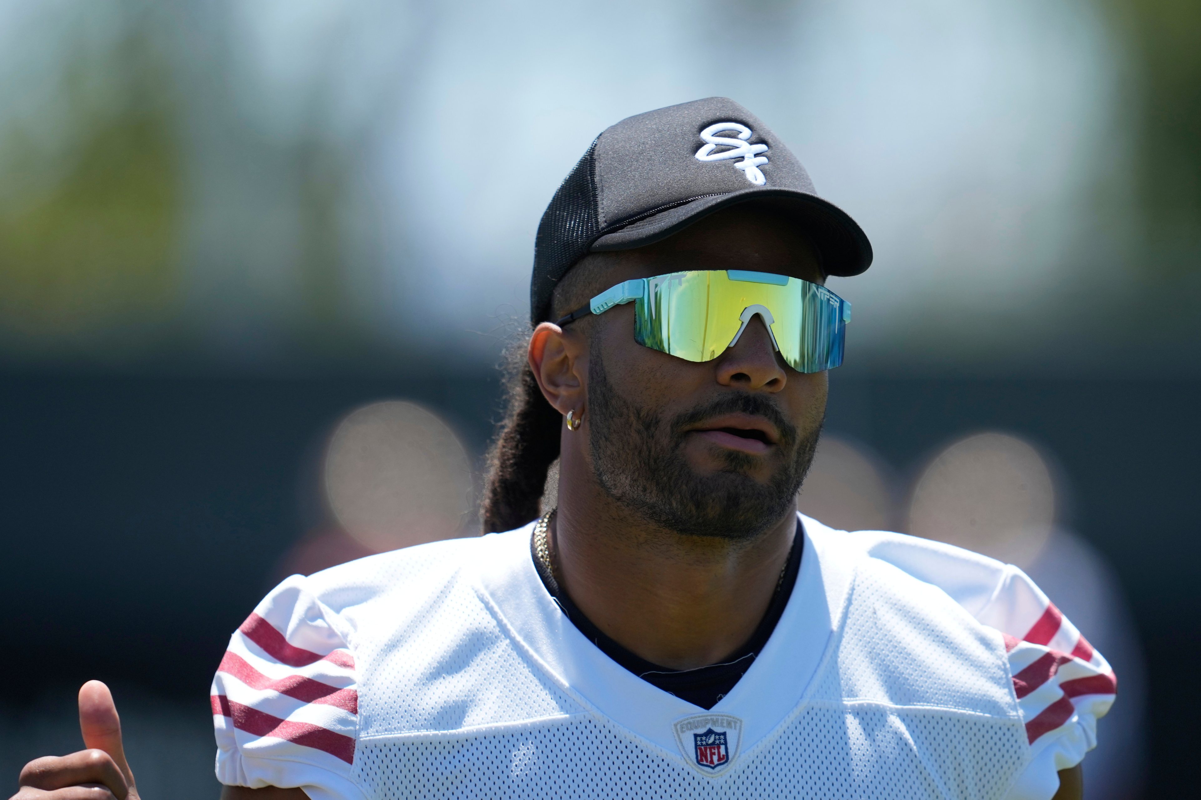 A person in a football jersey and mirrored sunglasses stands outdoors. They wear a black cap with white letters and have a small earring and facial hair.