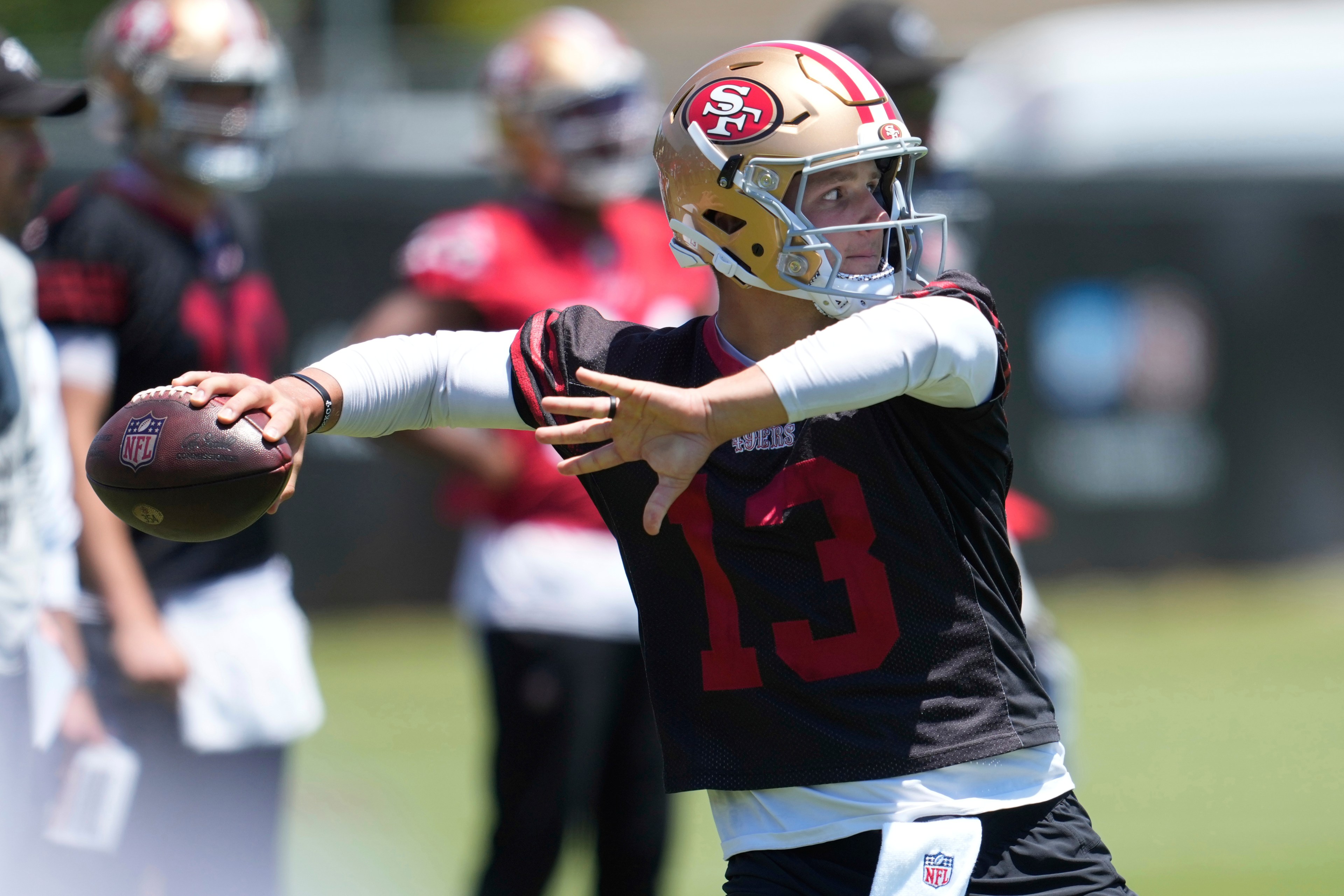 A football player wearing a gold helmet and black jersey prepares to throw a football. The number 13 is visible, and teammates are in the background.