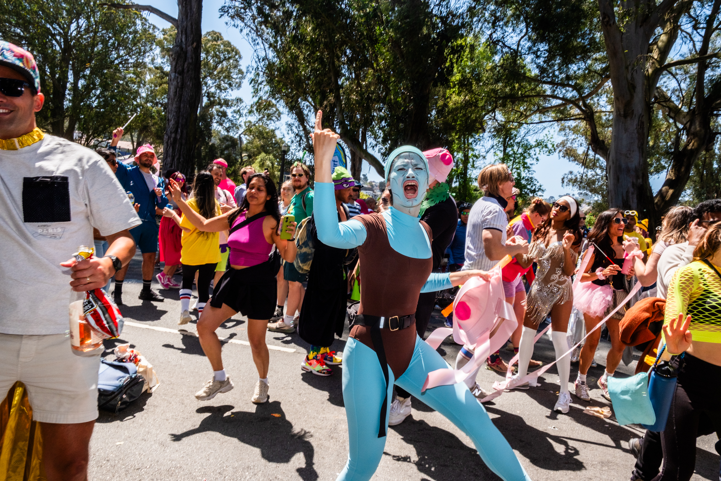 The image depicts a lively group in colorful, eccentric costumes at an outdoor event, with trees in the background. A person in blue is energetically posing.