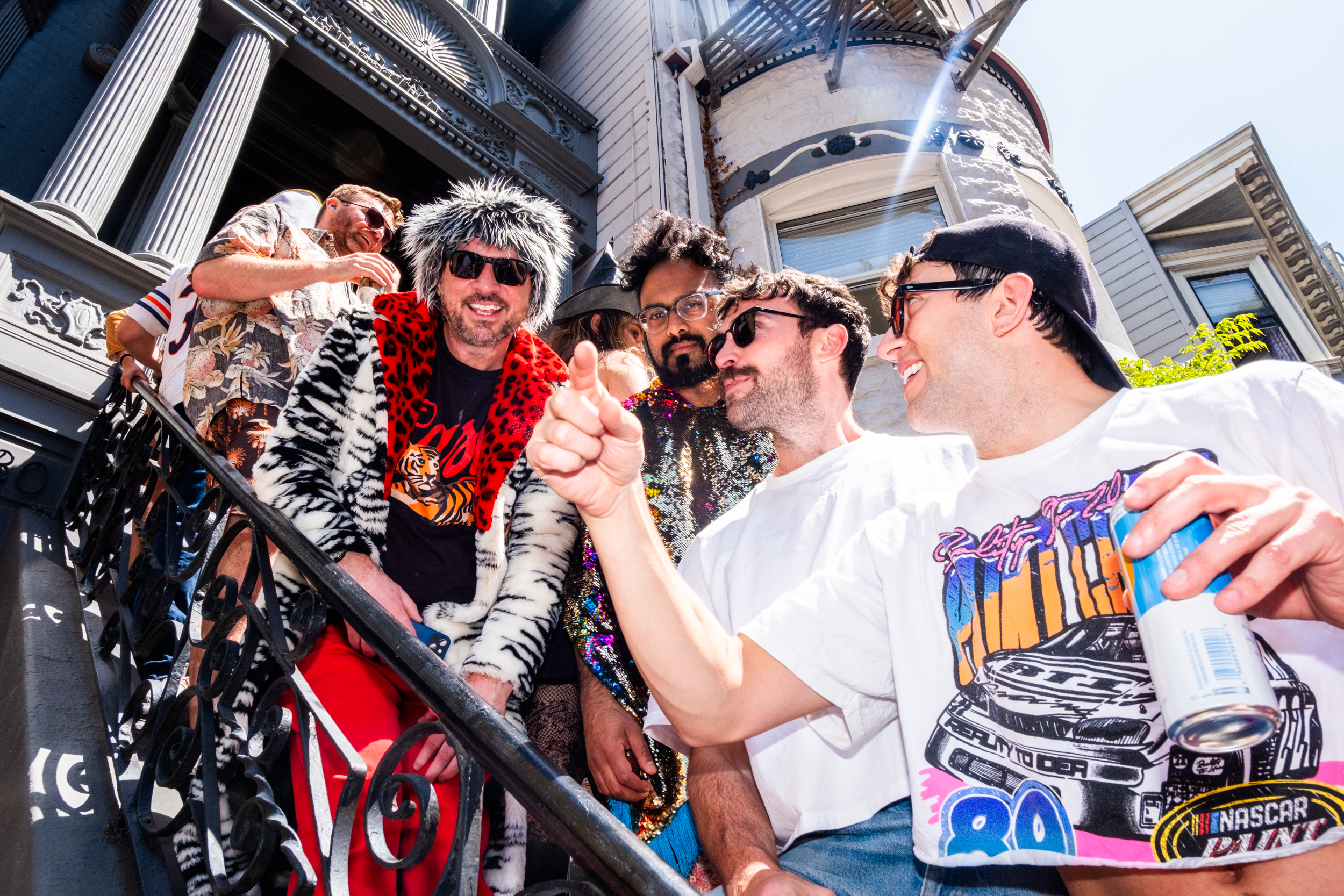 A group of five people stands on a staircase outside a building. They wear colorful, eccentric outfits, including fur and sequin tops, and are smiling and chatting.