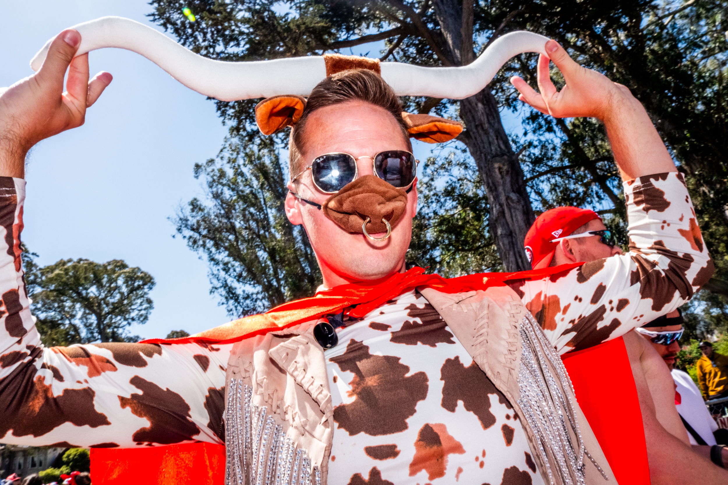 A person in a cow-themed costume with sunglasses, a nose ring, and large horns holds their hands up. They're outdoors with trees in the background.