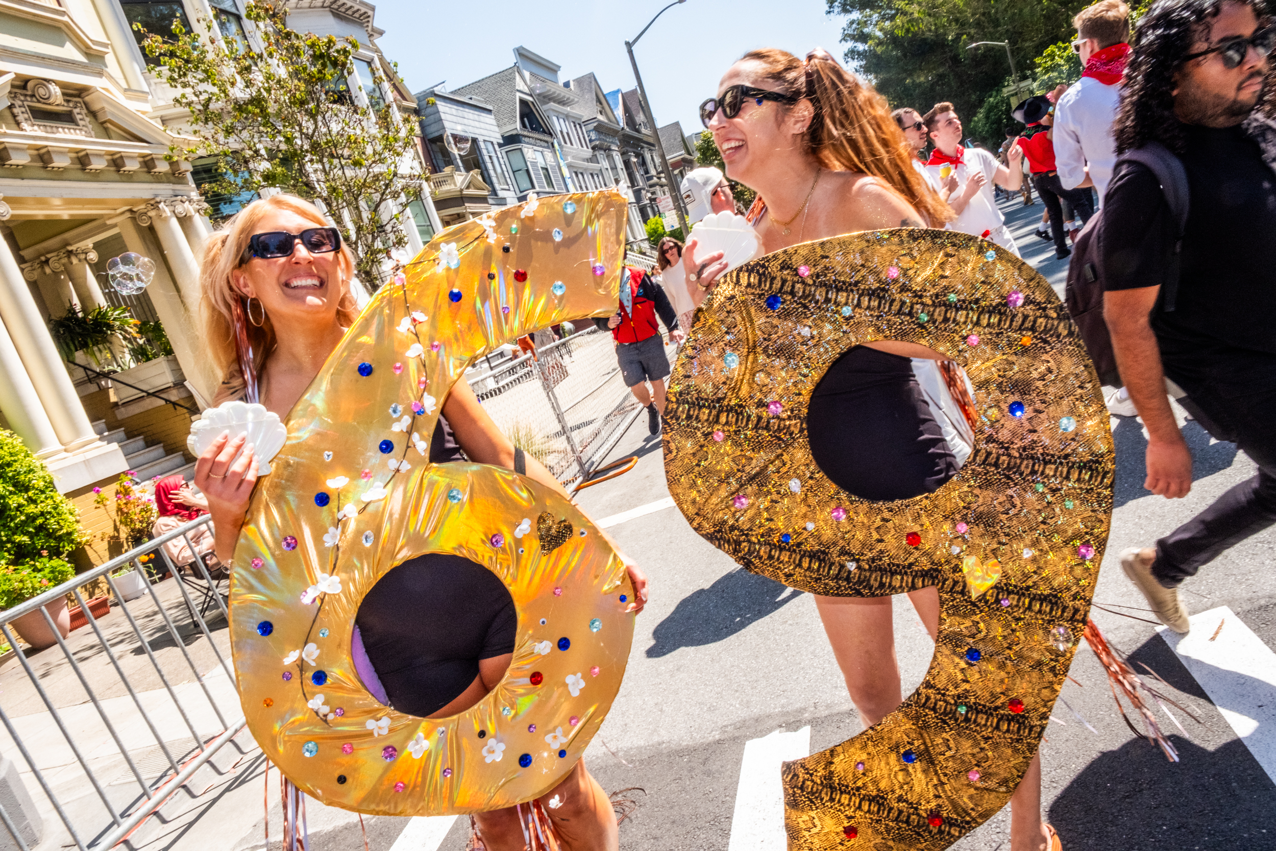 Two women walk outdoors holding large glittery numbers, 6 and 9, adorned with decorations. They smile and wear sunglasses, surrounded by people on a sunny day.