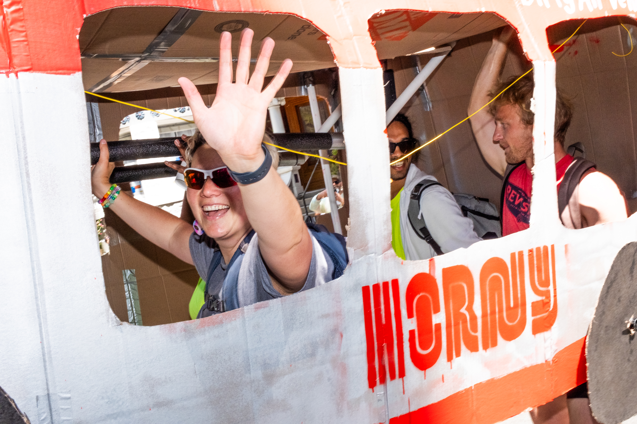 A person in sunglasses and a gray shirt waves enthusiastically from a cardboard structure resembling an airplane, painted white and red with visible cutouts.