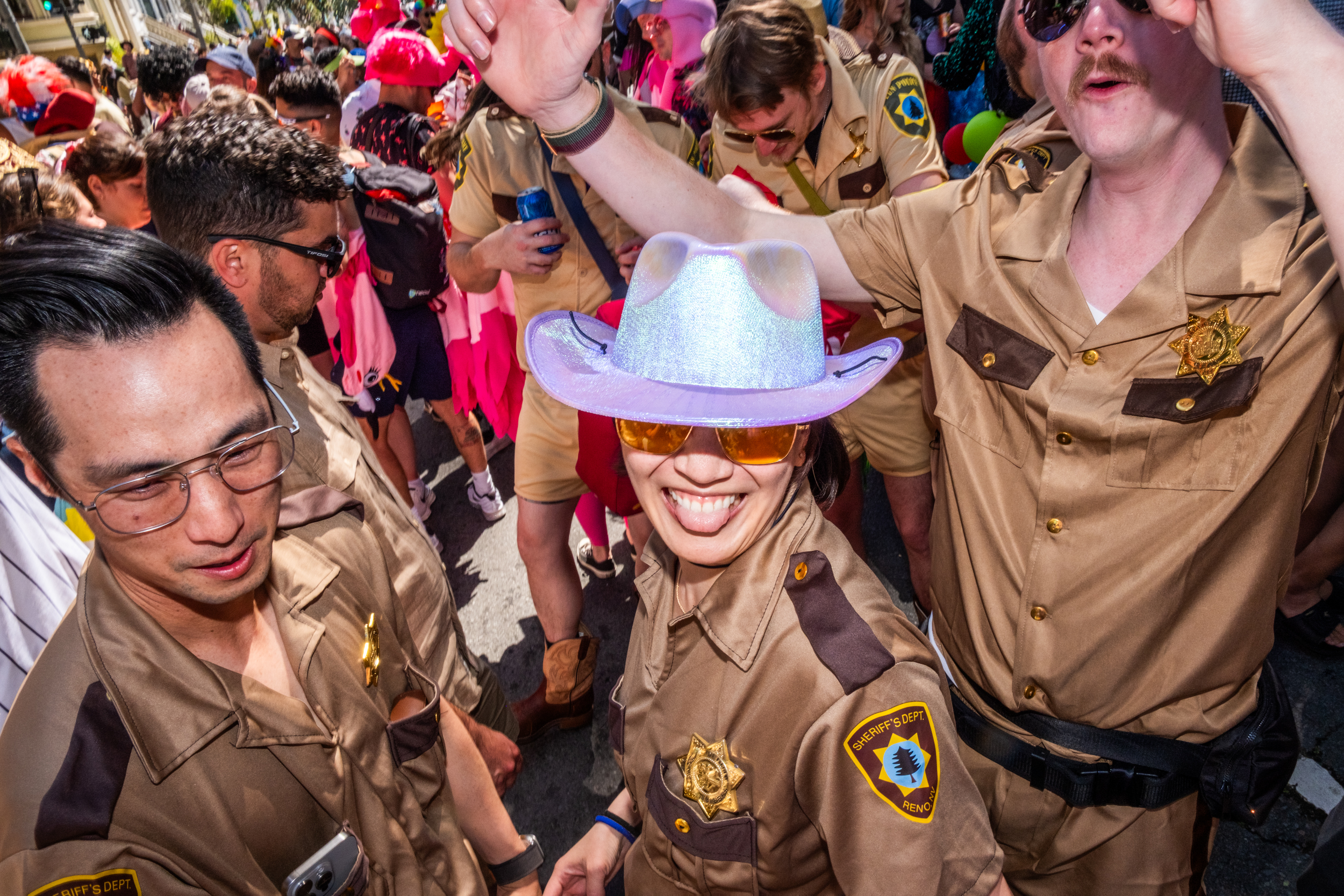 A group of people is dressed in tan sheriff uniforms with badges, smiling and interacting at a lively outdoor event. One wears a shiny cowboy hat.