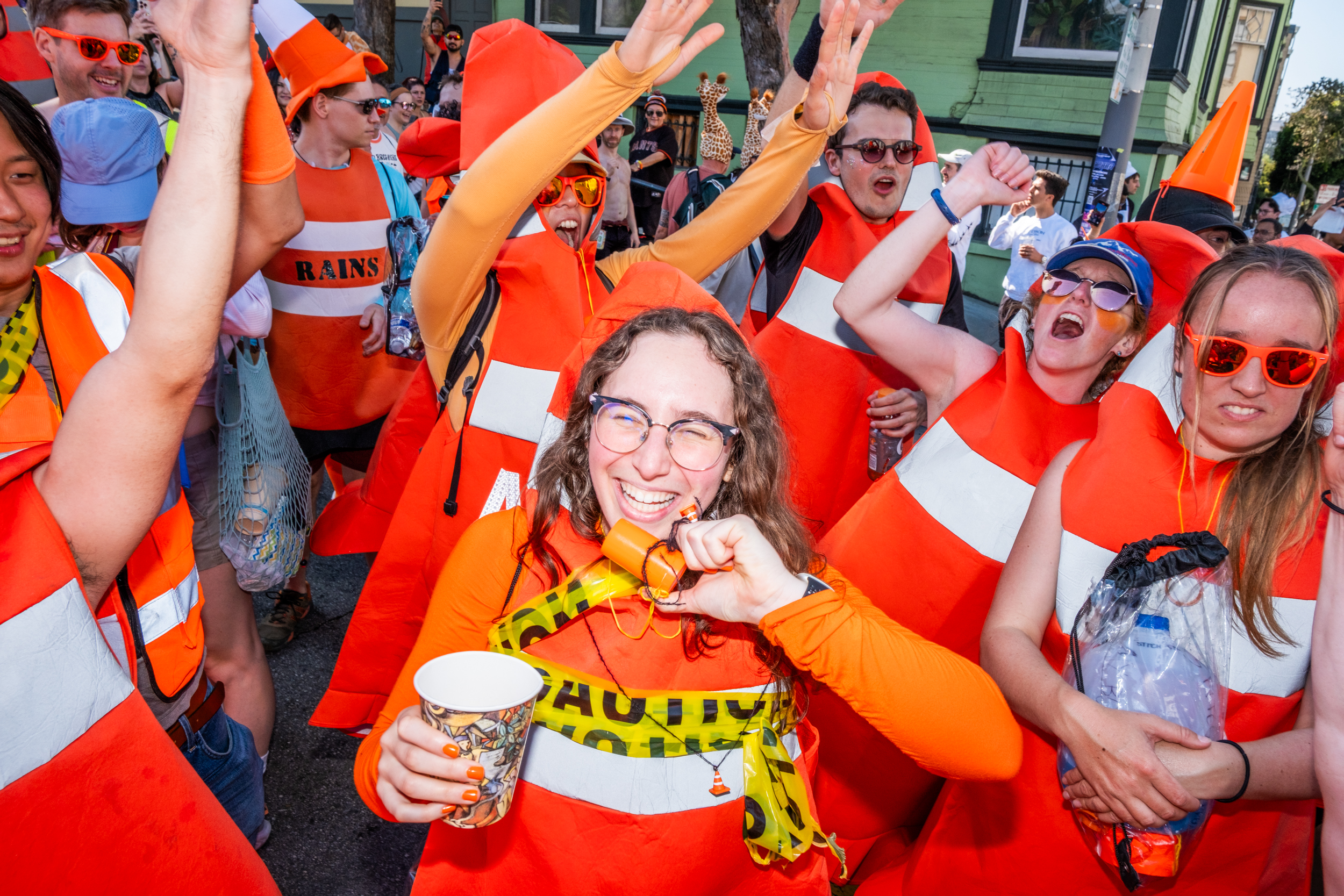 A group of people is celebrating, dressed as orange traffic cones. They are smiling and holding drinks, enjoying a lively outdoor event.
