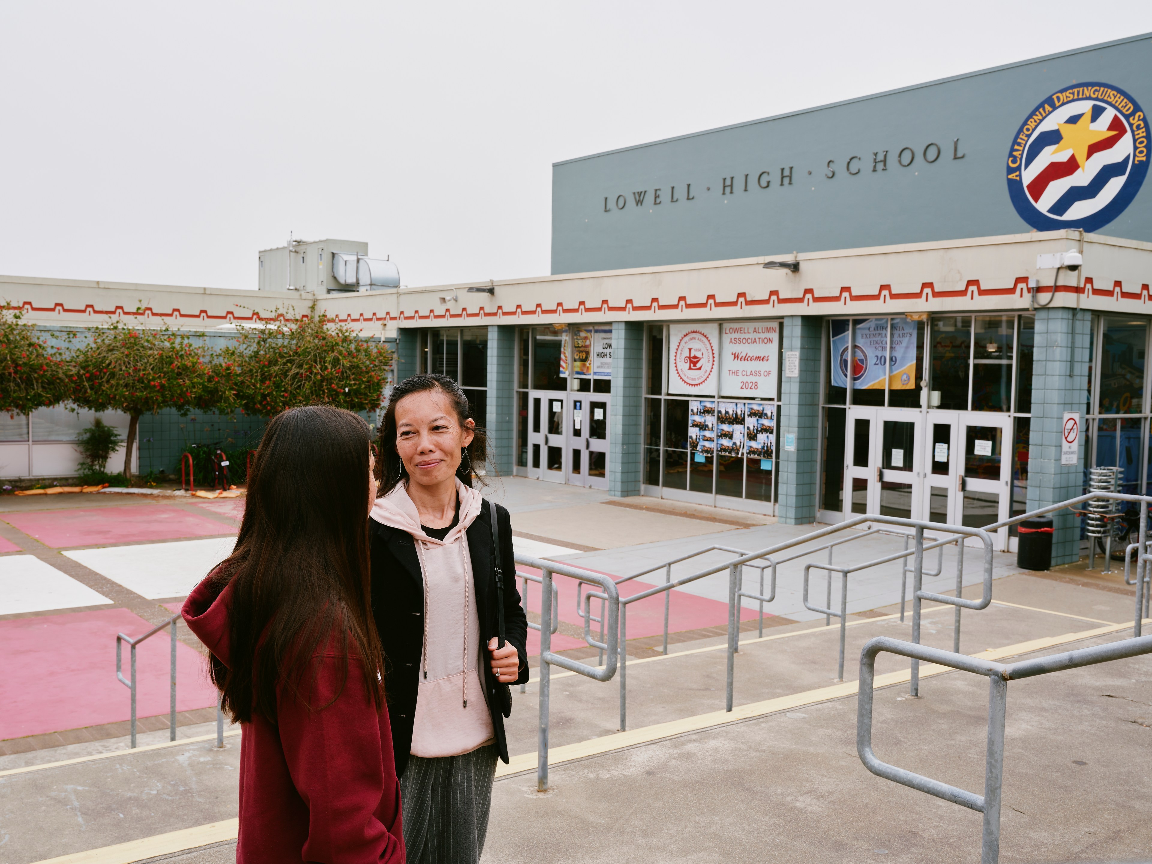 Two people stand outside Lowell High School. The building has a sign for a California Distinguished School and posters for the Class of 2028.