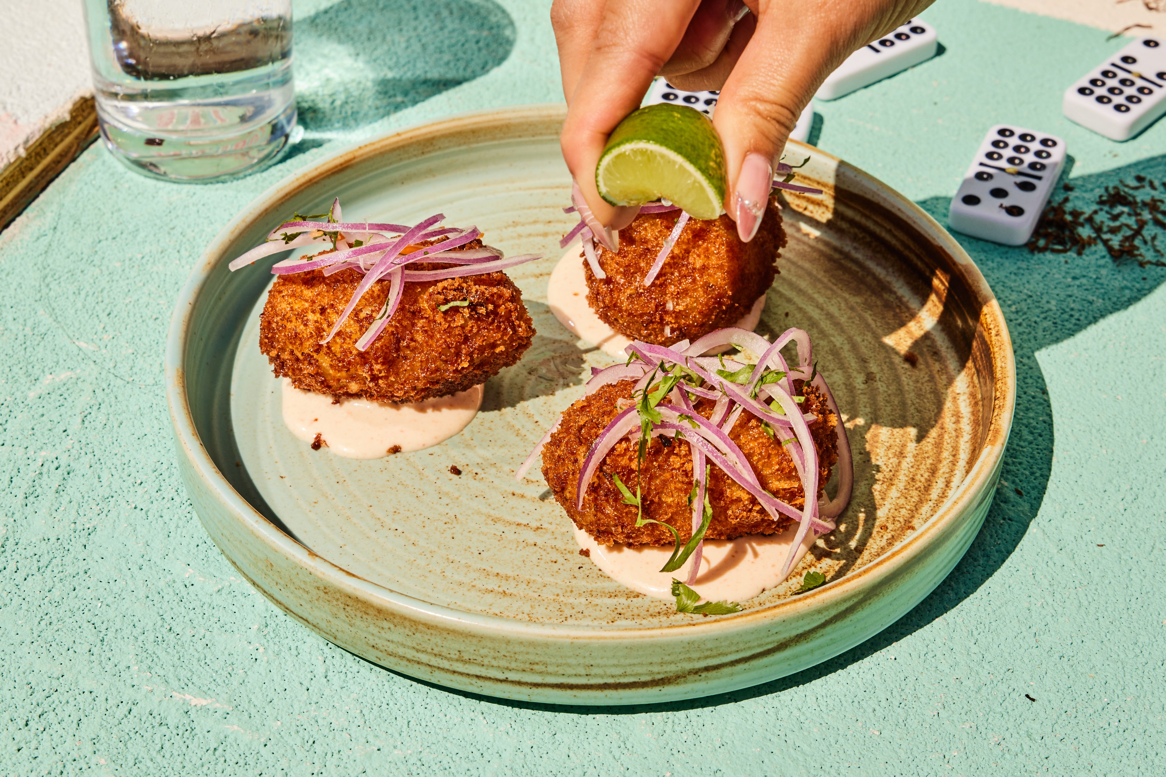 Three crispy croquettes sit on sauce in a round plate, garnished with purple onions and herbs. A hand squeezes lime over the dish, with dominoes nearby.