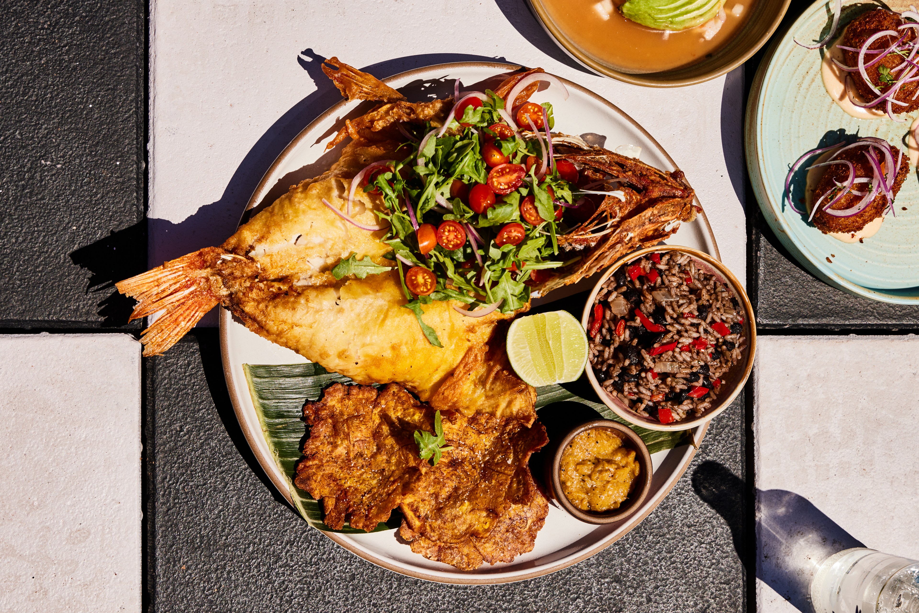 The image shows a plate with fried fish topped with a fresh salad of greens and cherry tomatoes, served with rice and beans, a lime wedge, and a small bowl of sauce.