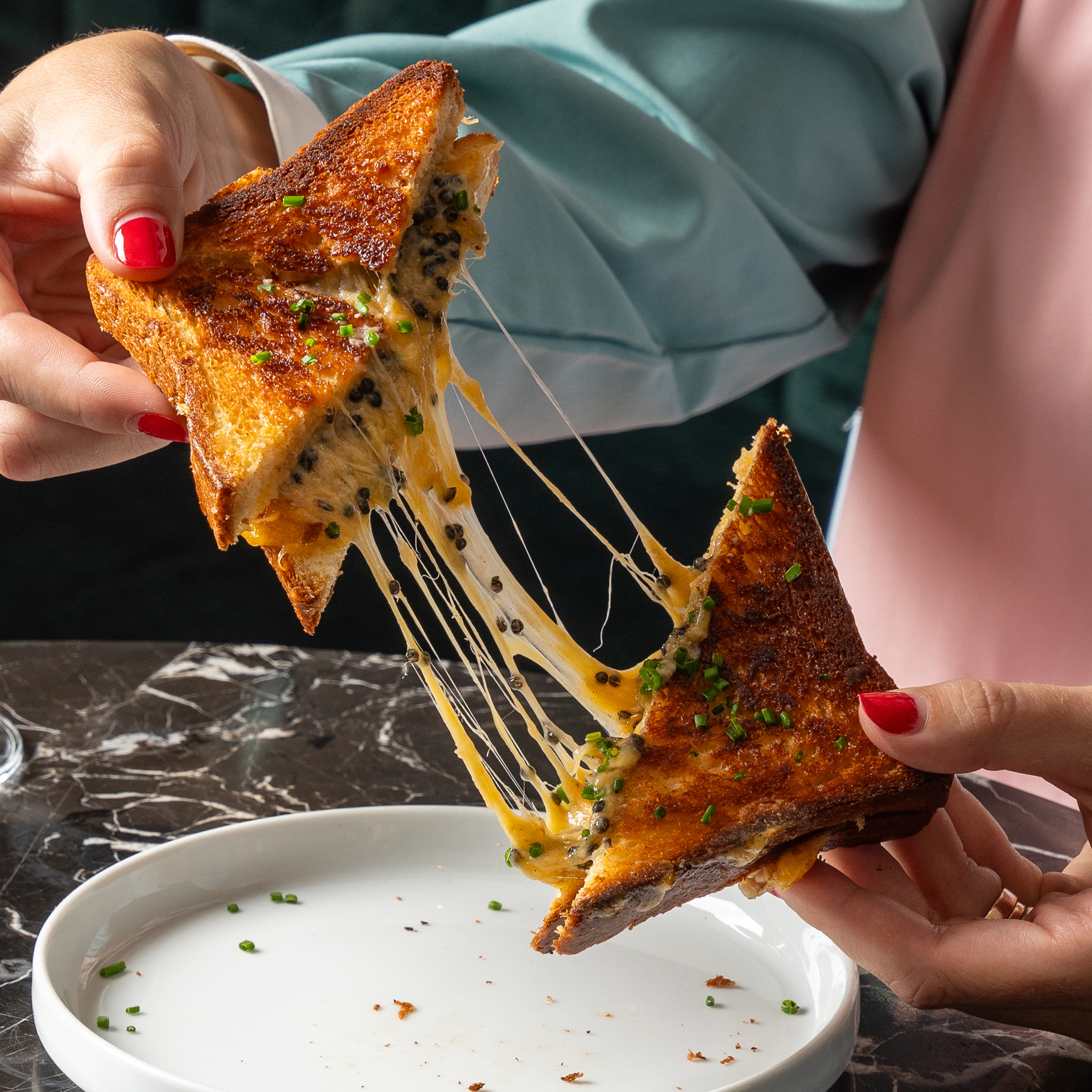 A person pulls apart a grilled cheese sandwich, revealing strings of gooey cheese. The sandwich is topped with herbs and rests over a white plate.