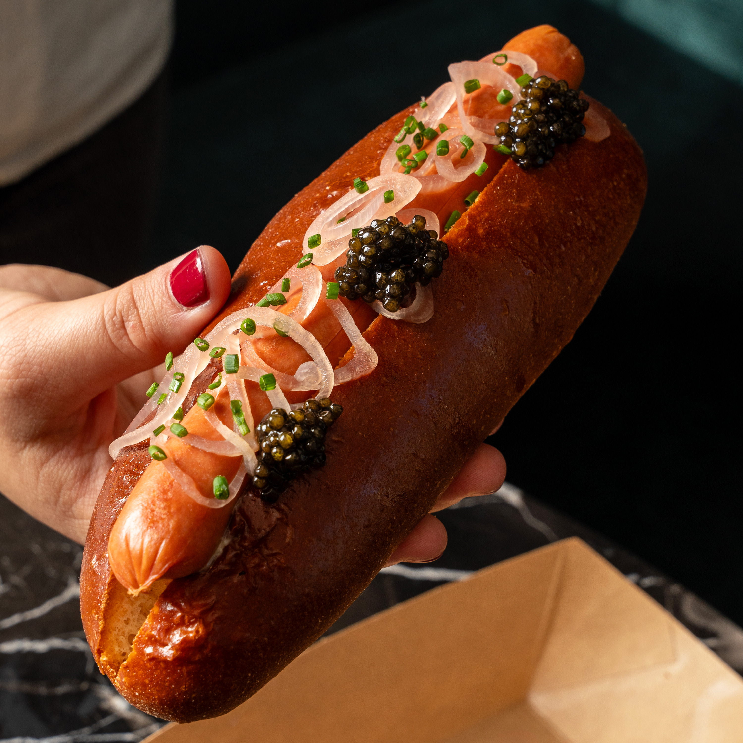 A person holds a gourmet hot dog in a shiny bun, topped with caviar, thin onion slices, and chopped chives, with a box in the background.