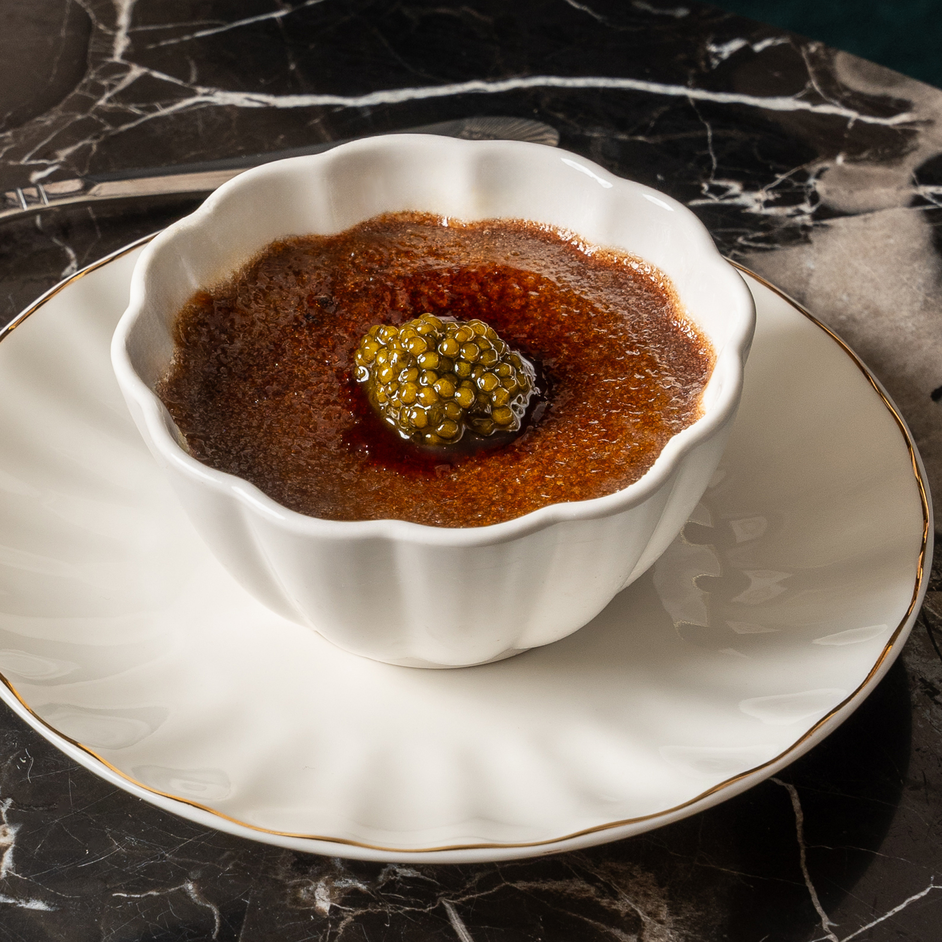 A white fluted bowl holds crème brûlée topped with caviar, sitting on a matching plate with a gold rim, placed on a dark, marbled surface.