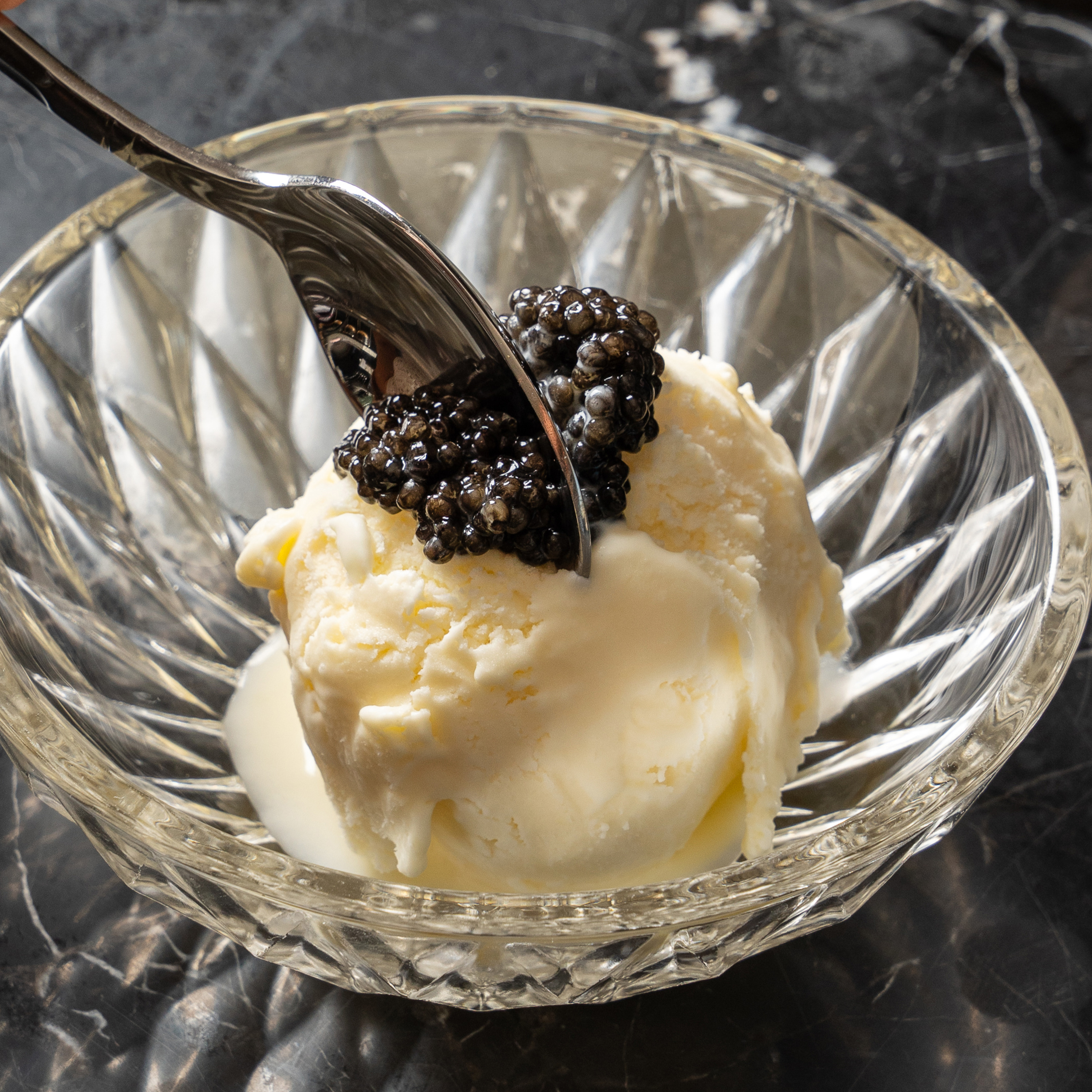 A scoop of vanilla ice cream is topped with black caviar in a faceted glass bowl, with a metal spoon ready to scoop some up.