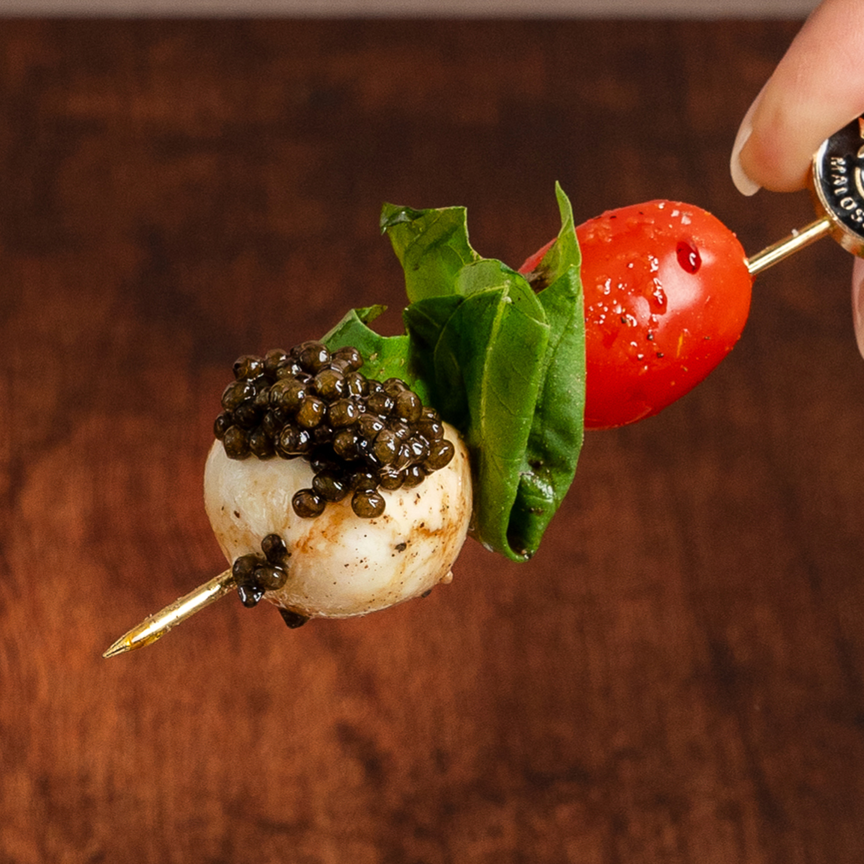 A skewer holds a cherry tomato, basil leaf, and mozzarella ball topped with caviar, set against a wooden background.