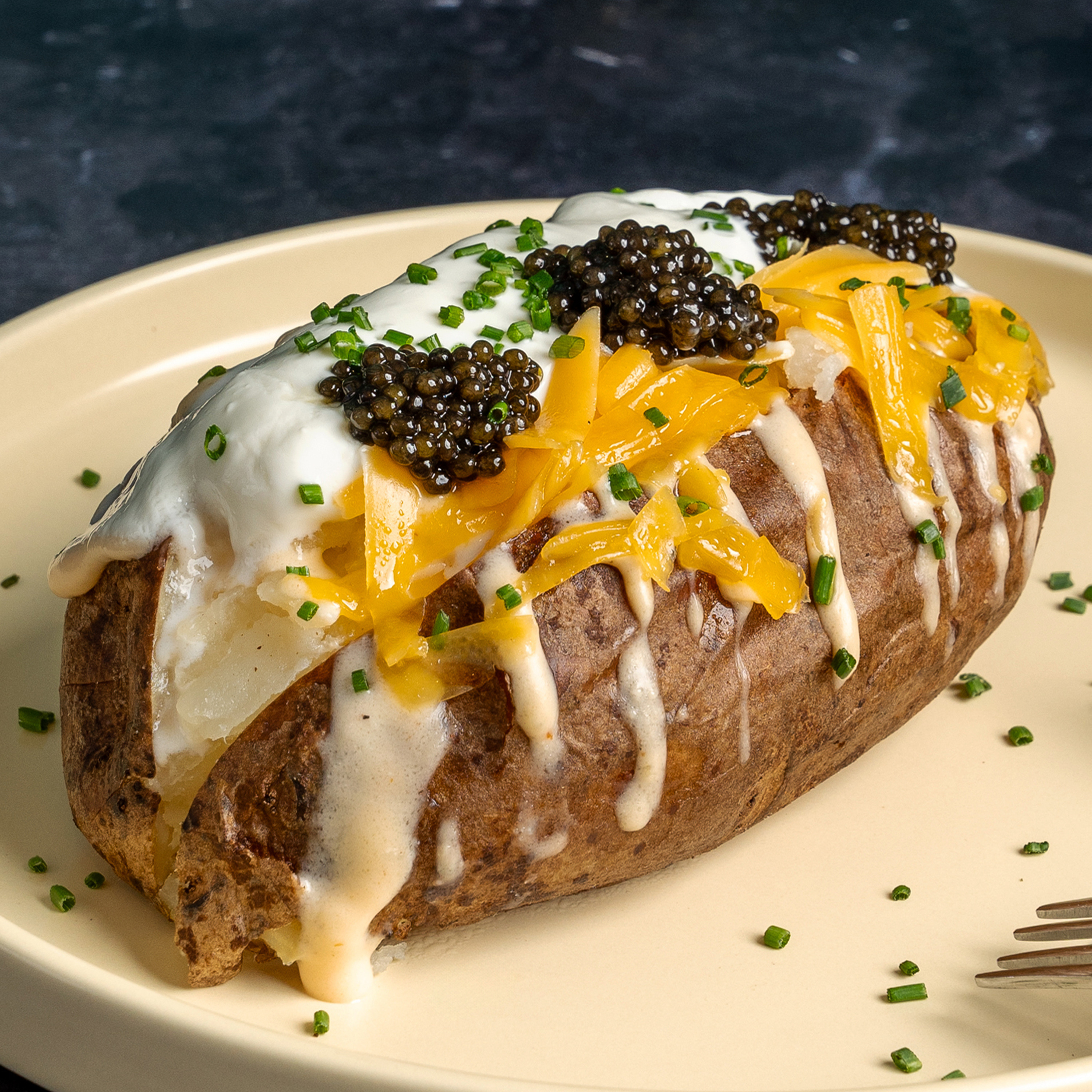 A baked potato topped with sour cream, cheddar cheese, black caviar, and chopped chives, sits on a beige plate, with a fork beside it.