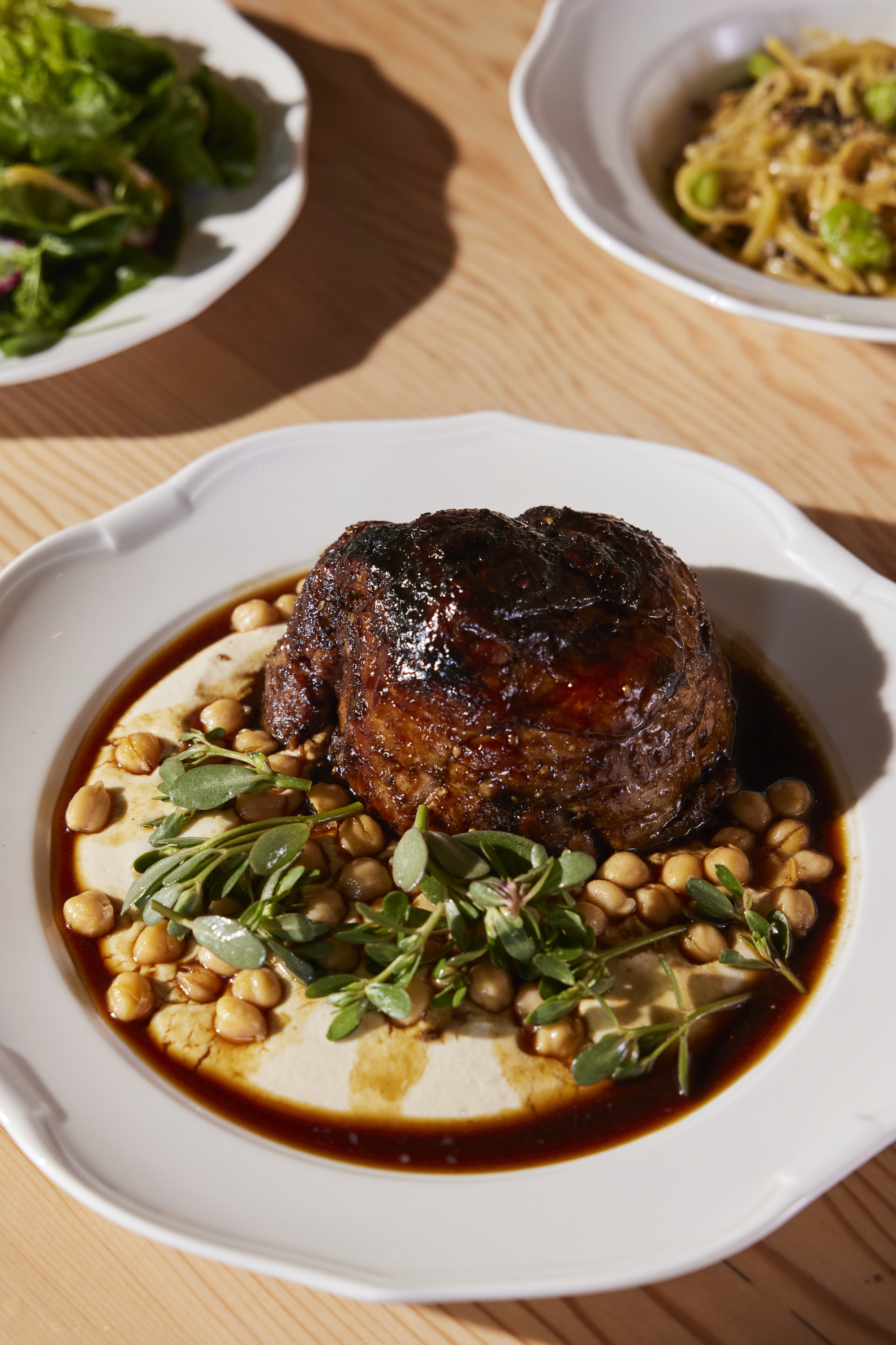 A glazed piece of meat sits on a plate with chickpeas, sauce, and garnished greens. Nearby dishes include a leafy salad and pasta with green peas.