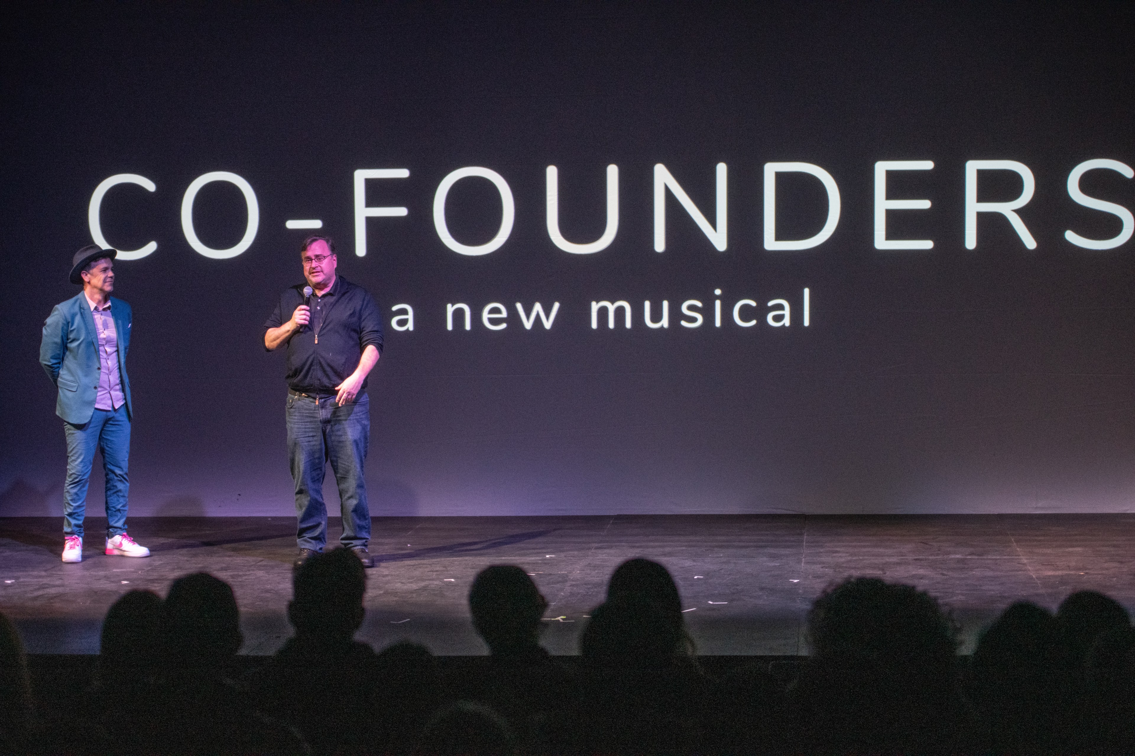 Two people stand on a stage in front of an audience. A large screen behind them displays "CO-FOUNDERS a new musical." One holds a microphone.
