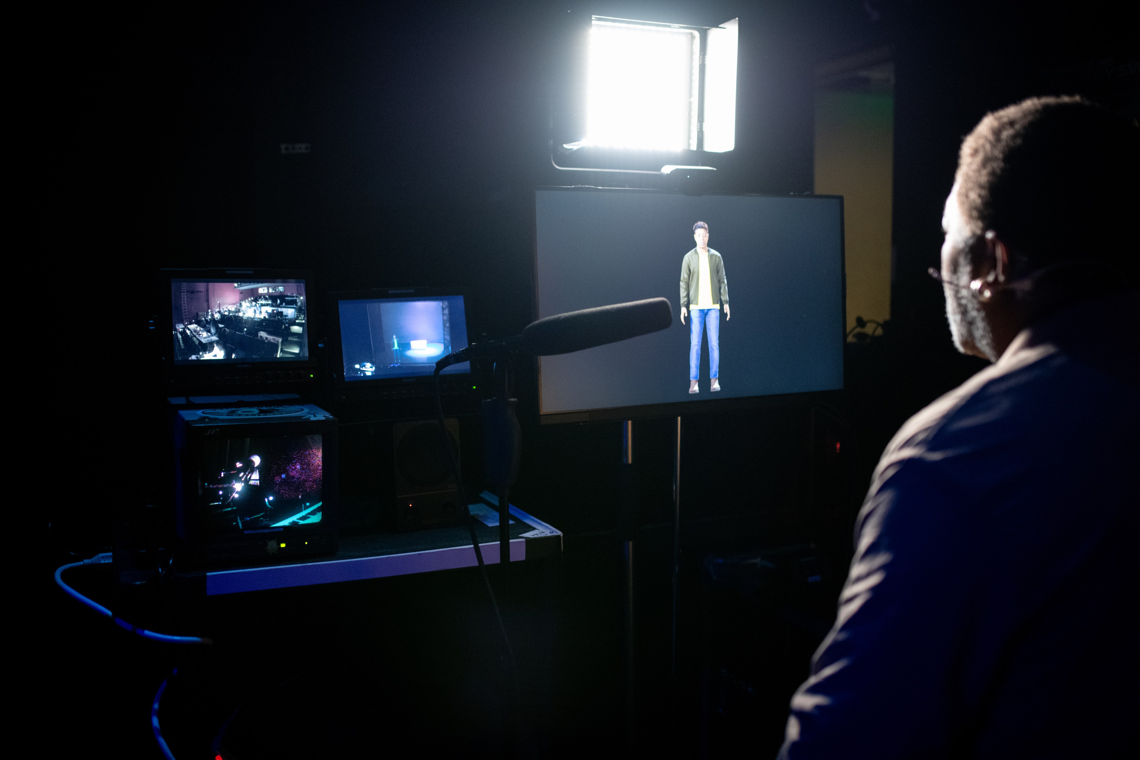 A person is facing a monitor displaying a virtual character. There are small screens on the side showing various video feeds, and a microphone is visible.