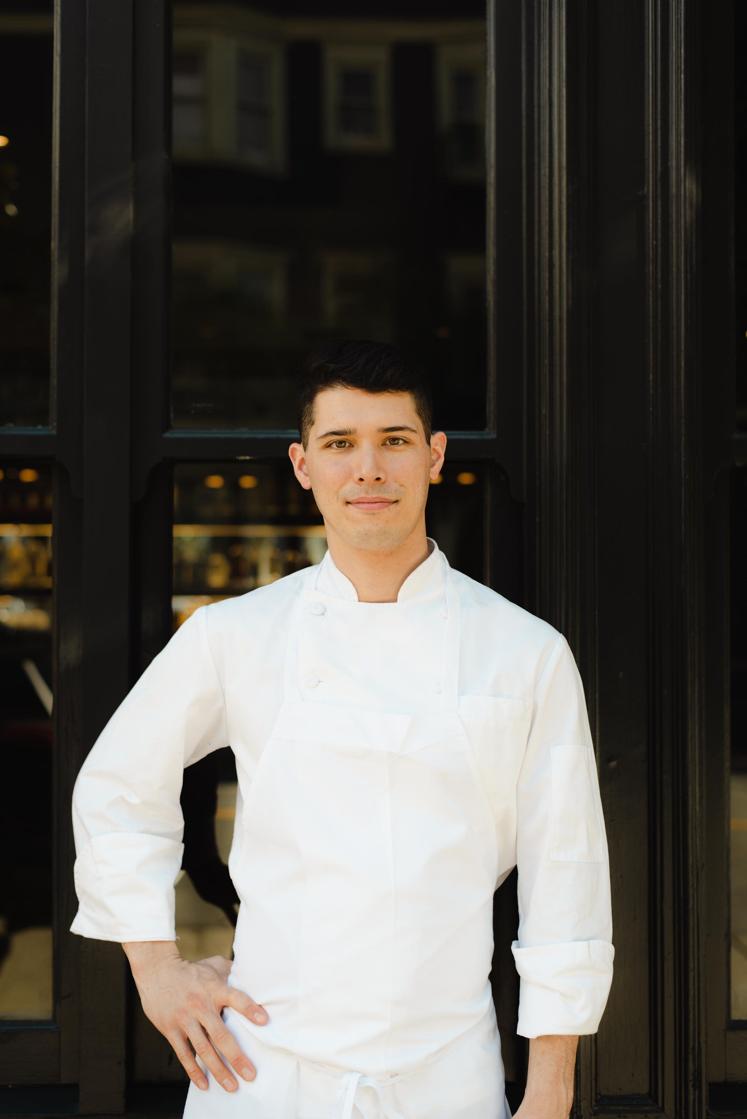 A person in a white chef's uniform stands confidently with hands on hips in front of a dark-framed window.