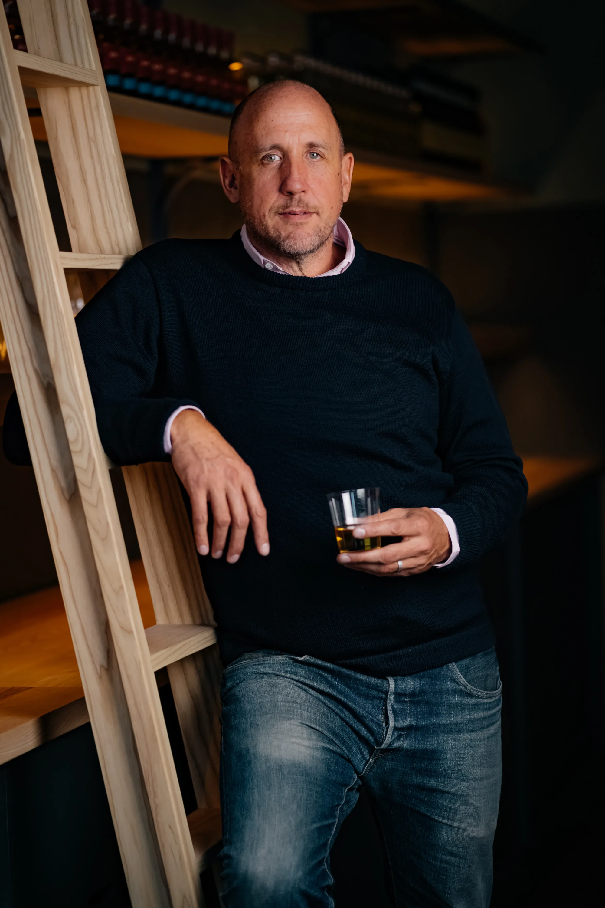 A man leans casually against a wooden ladder, holding a glass of amber liquid. He's wearing a dark sweater and jeans, in a warmly lit setting.