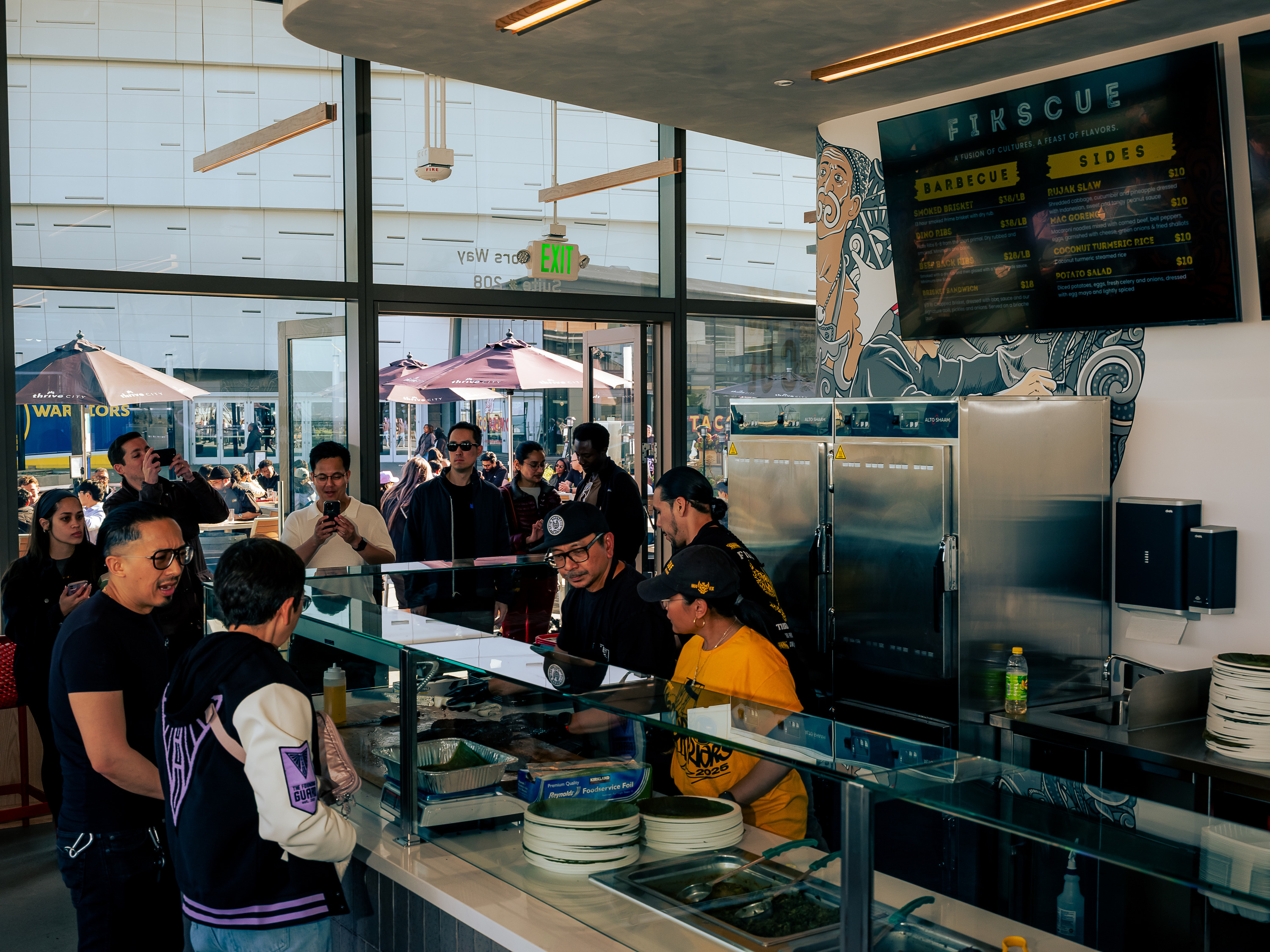 People are gathered inside a restaurant with a menu board above. Staff serve from behind a glass counter. Outside, more people are visible near umbrellas.