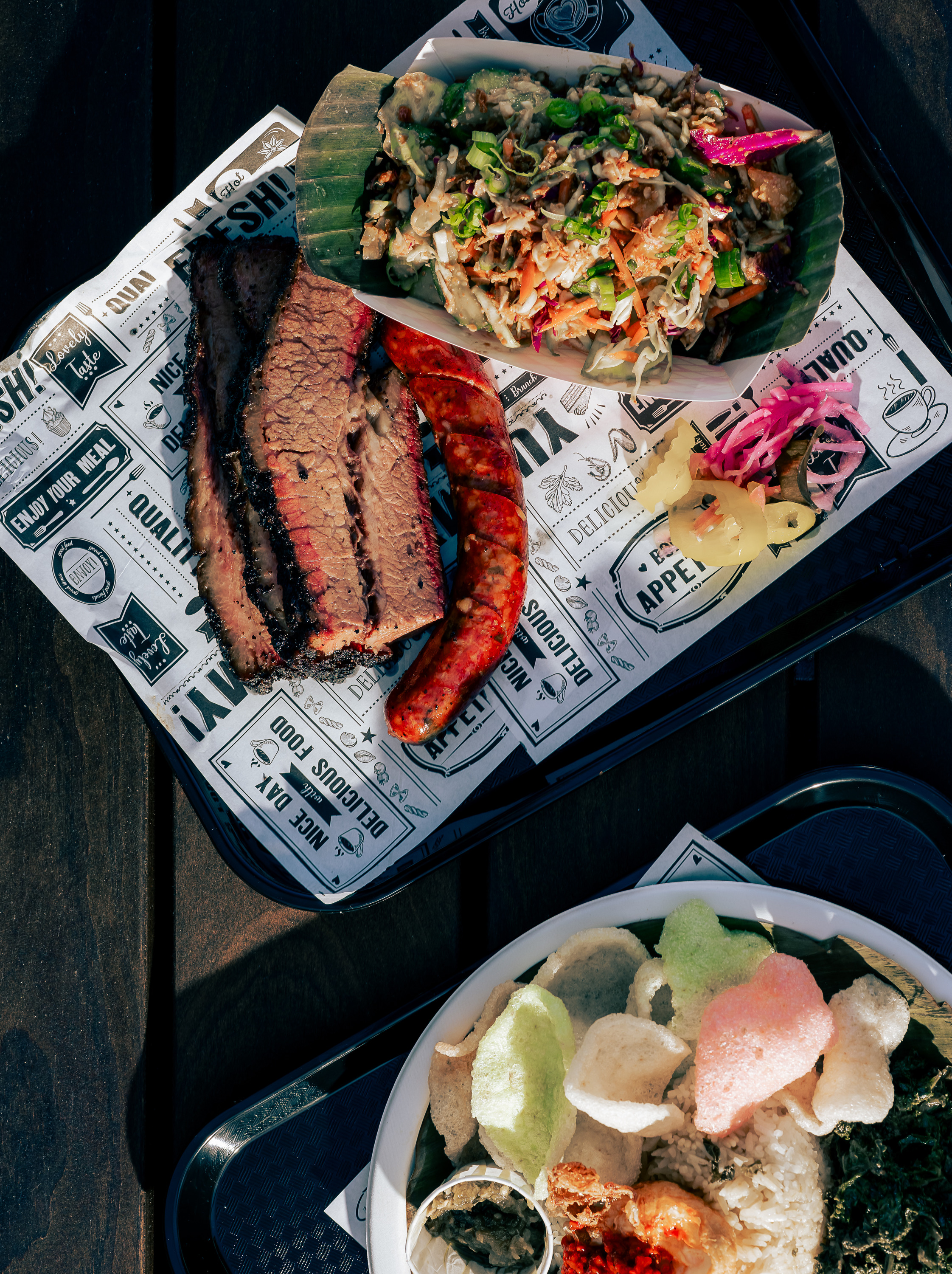 The image shows two plates of food. One has sliced brisket, sausage, and salad on decorative paper, and the other has rice, chips, greens, and a condiment.
