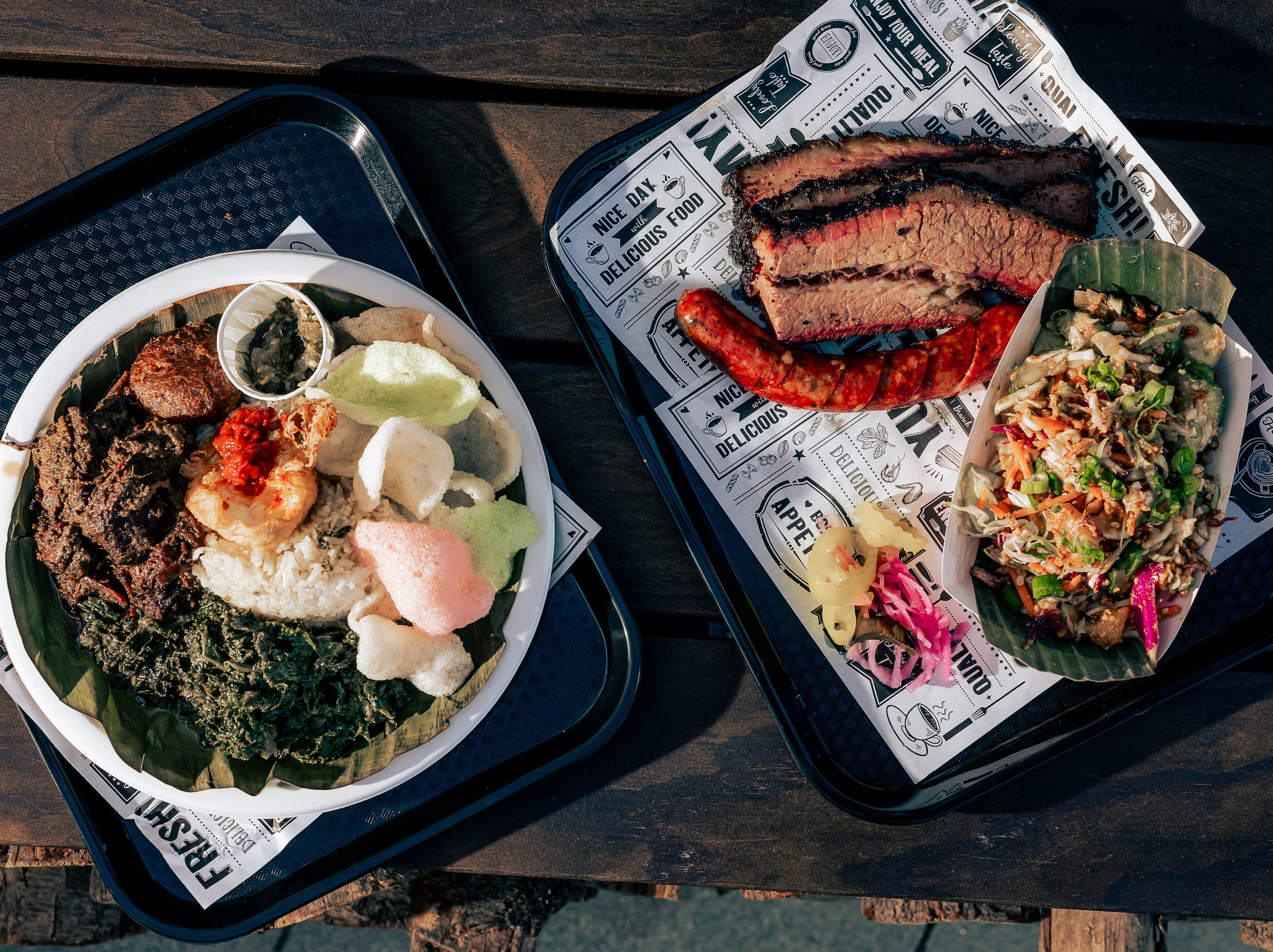 The image shows two trays of food: one with rice, meat, greens, crackers, and sauce, and the other with sliced brisket, sausage, pickles, and a vegetable salad.
