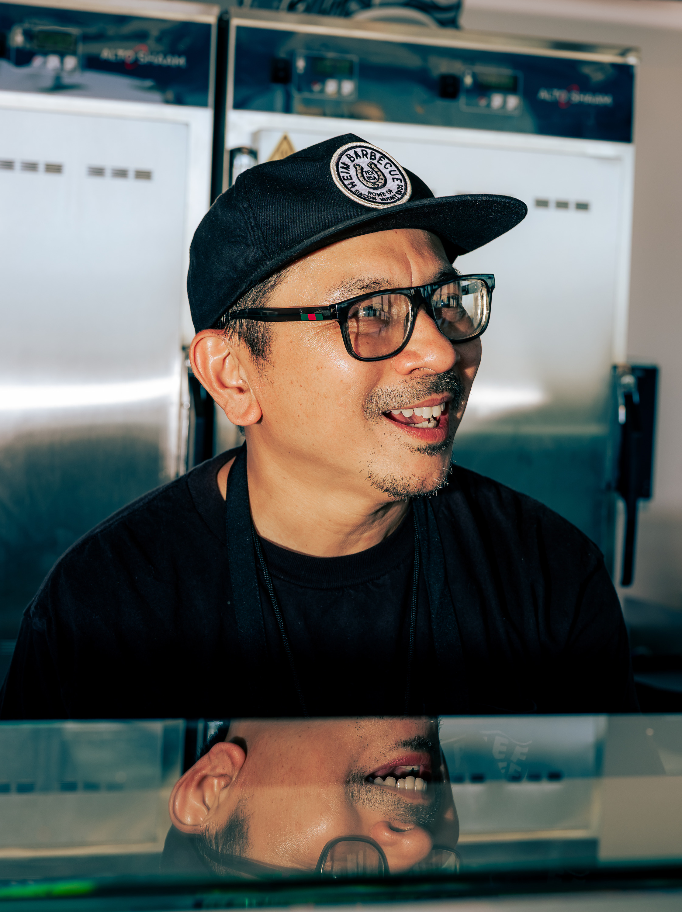 A man in glasses and a black cap with a patch is smiling. Behind him are metal appliances, and his reflection is visible on a glass surface.