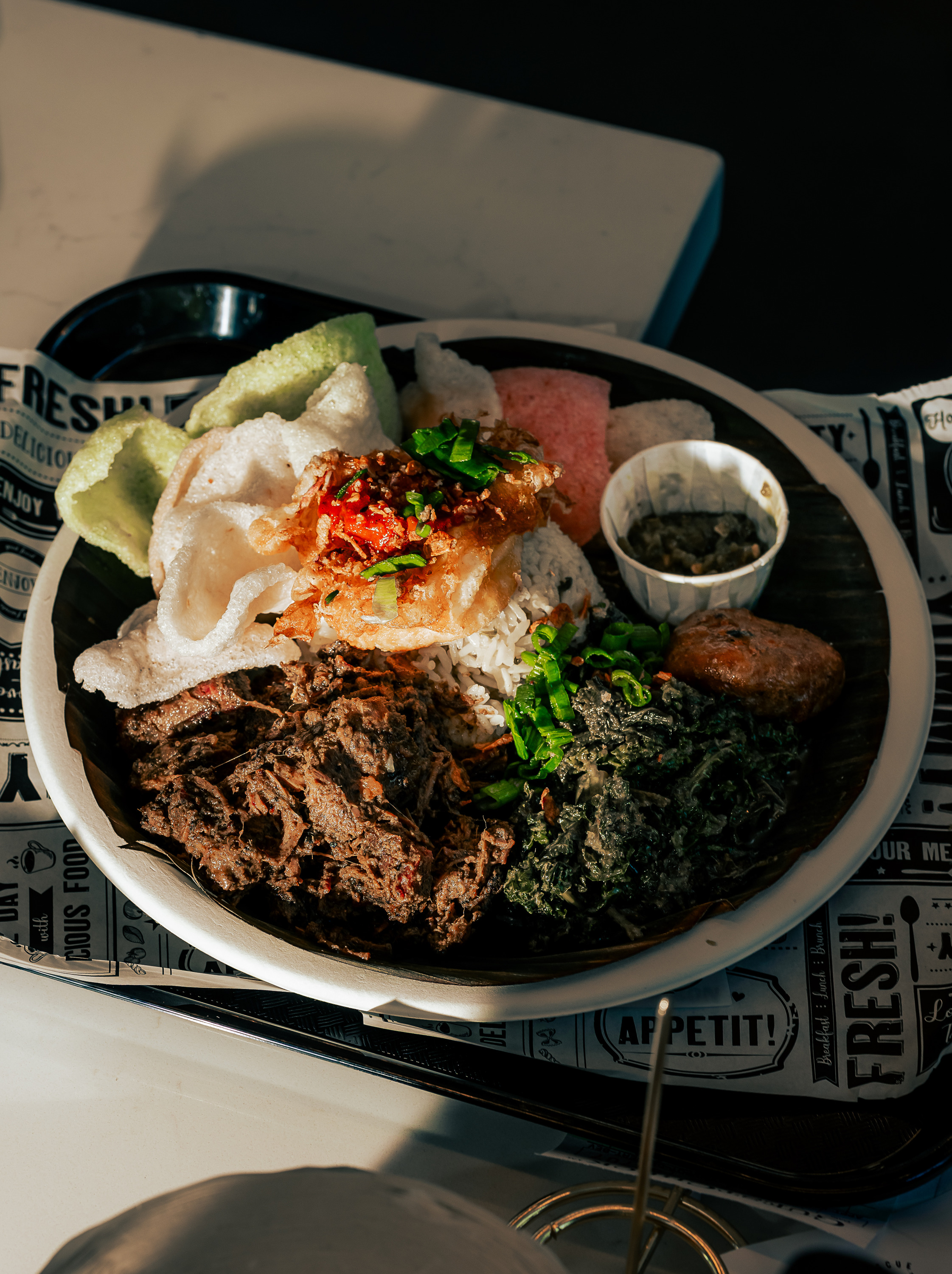 The image shows a plate of rice topped with shredded meat, greens, colorful prawn crackers, chili, and a small cup of sauce, set on printed paper.