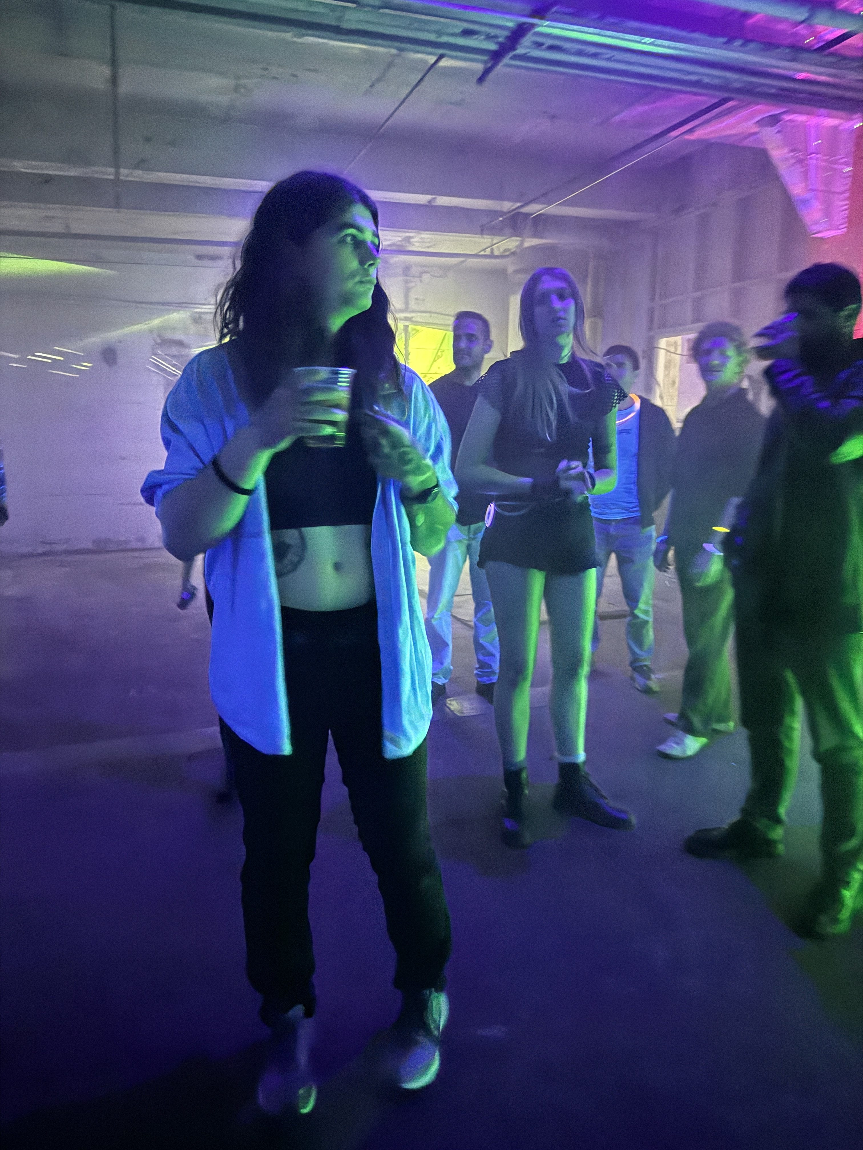A woman in casual attire holds a drink at a dimly lit event. Neon lights cast colorful glows on people standing and mingling in the background.