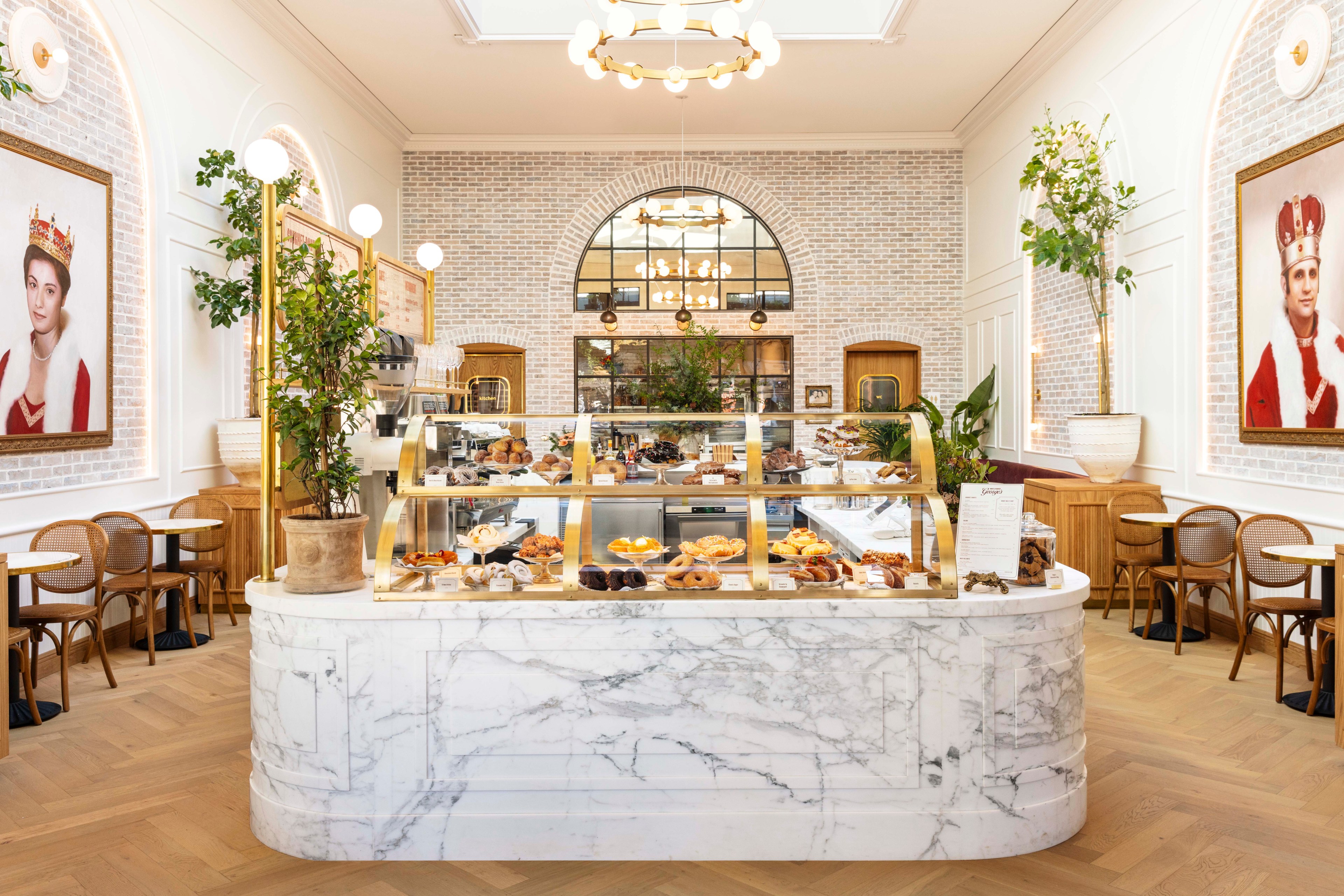 The image shows a bright bakery with a marble counter and glass display filled with pastries. Elegant seating, indoor plants, and large portraits adorn the space.