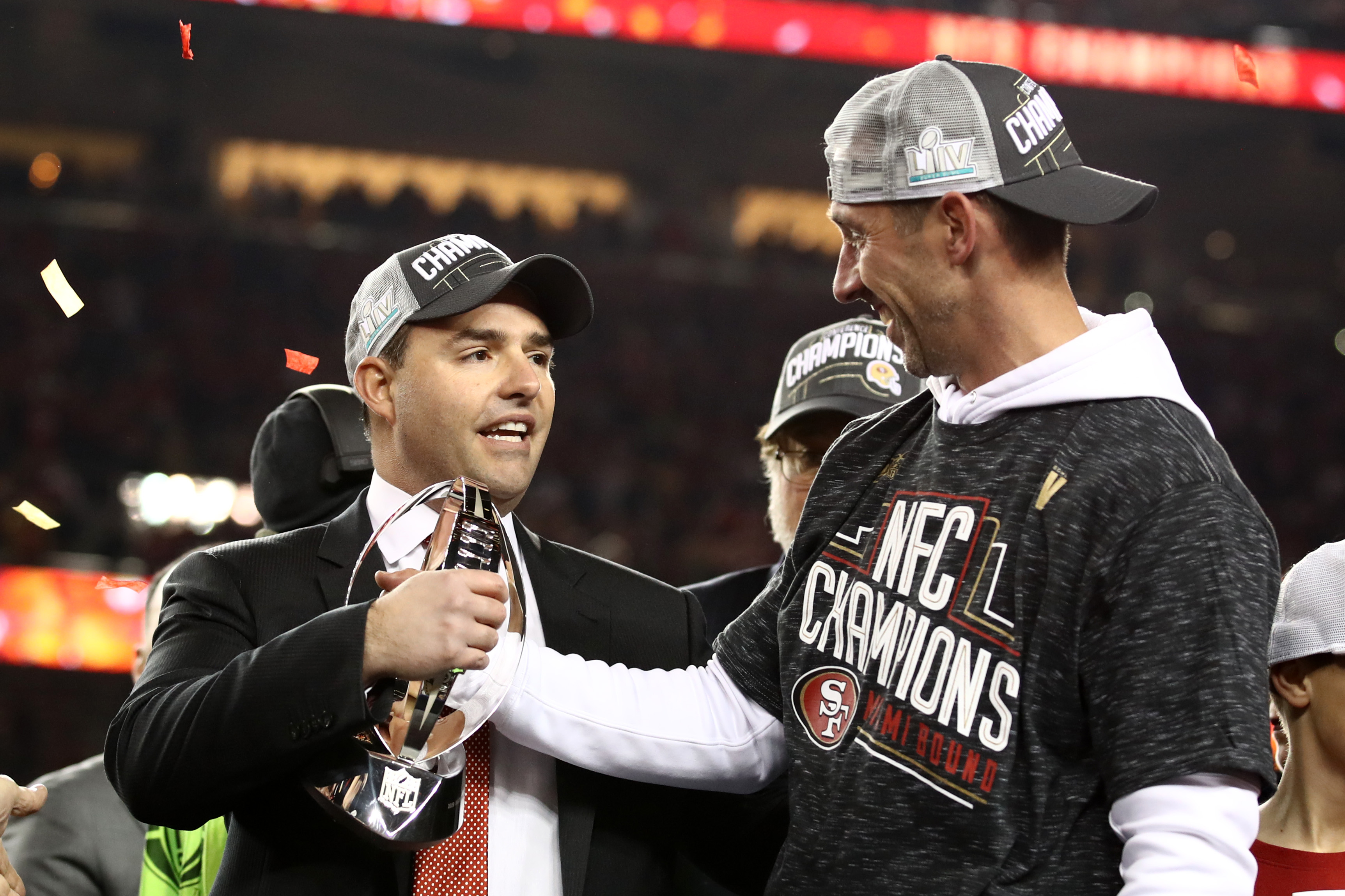 Two men wearing &quot;Champions&quot; caps celebrate with a trophy amid falling confetti. One wears a suit, the other an &quot;NFC Champions&quot; shirt.