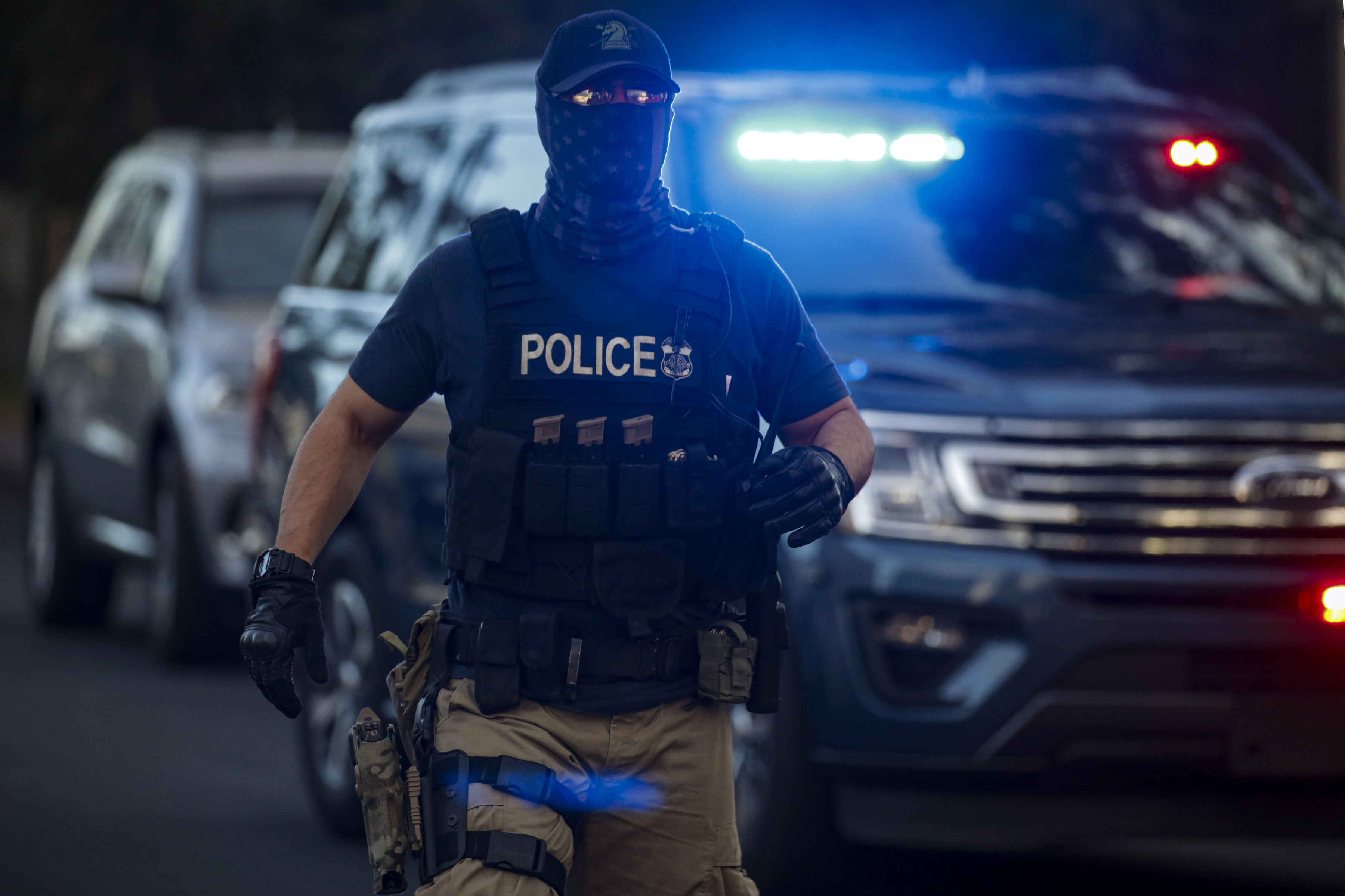 A police officer in tactical gear and face covering stands in front of a police vehicle with flashing lights. The scene suggests an active situation.