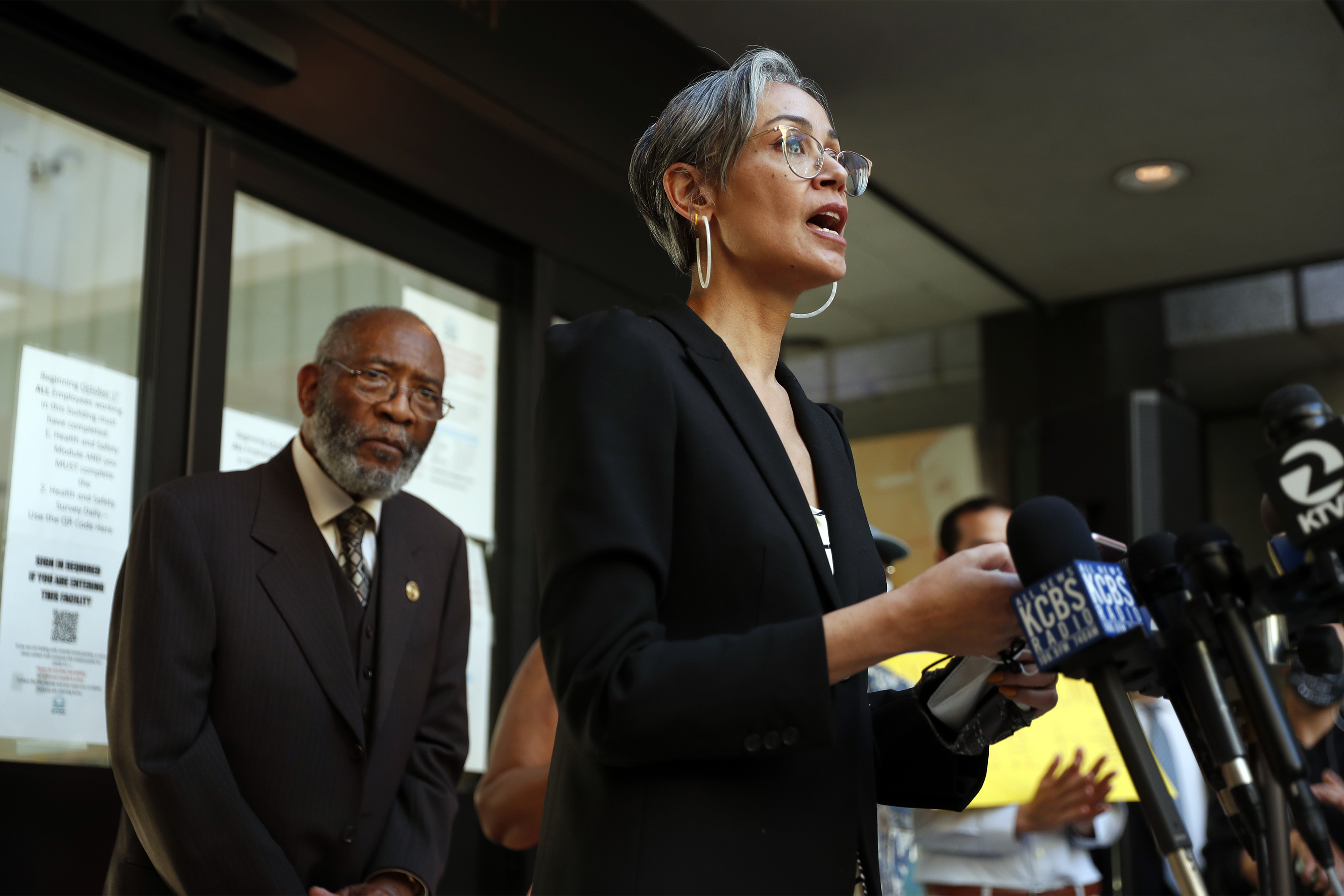 A person with short gray hair and glasses speaks at a podium surrounded by microphones, while an older gentleman in a suit stands behind them.